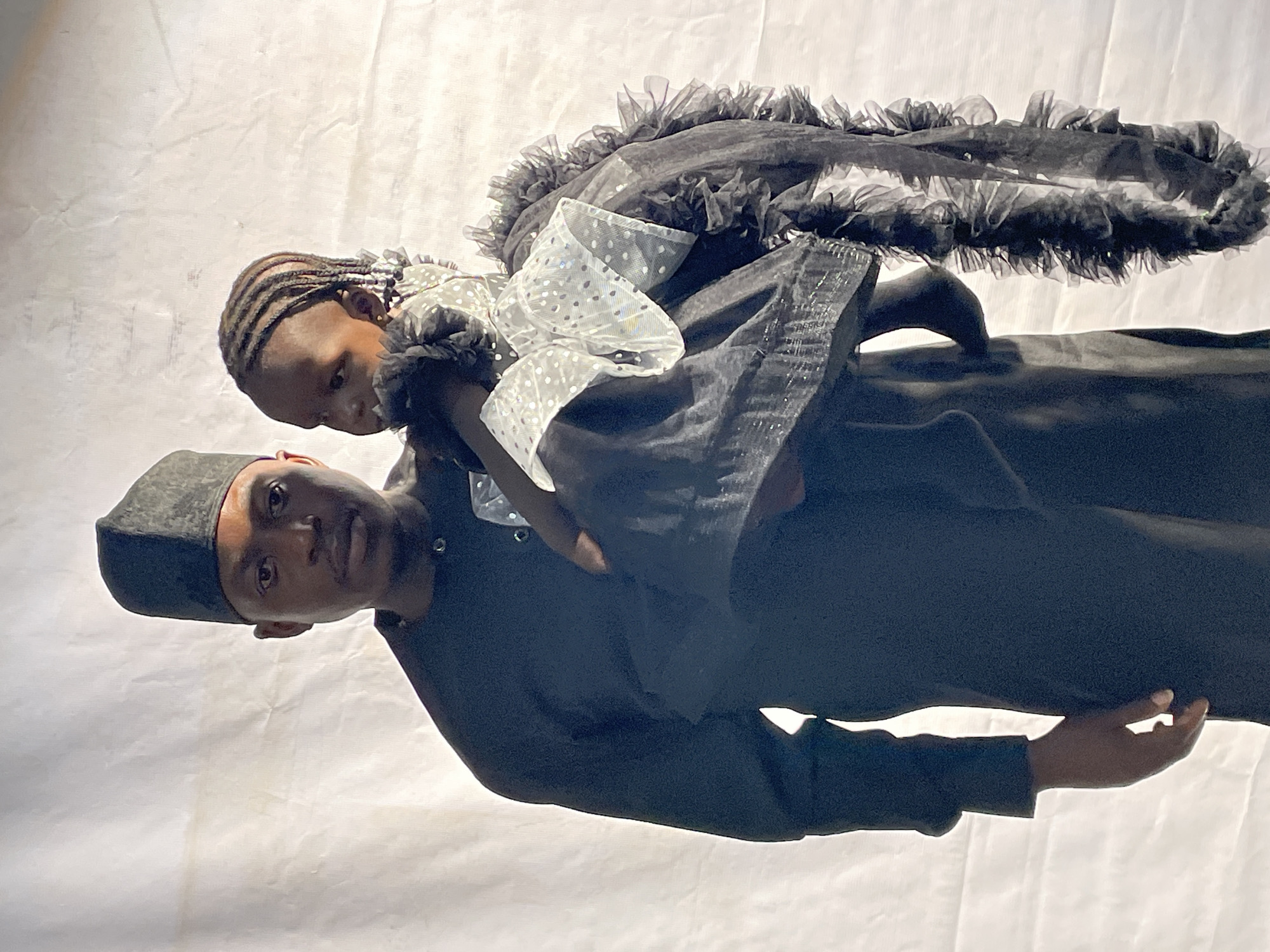 A man wearing a kufi hat and black clothing holds a child dressed in a black and white tulle dress. The background is a mottled white surface. The photo does not contain enough information to determine if it was taken in Barakin Akawo, Nigeria.