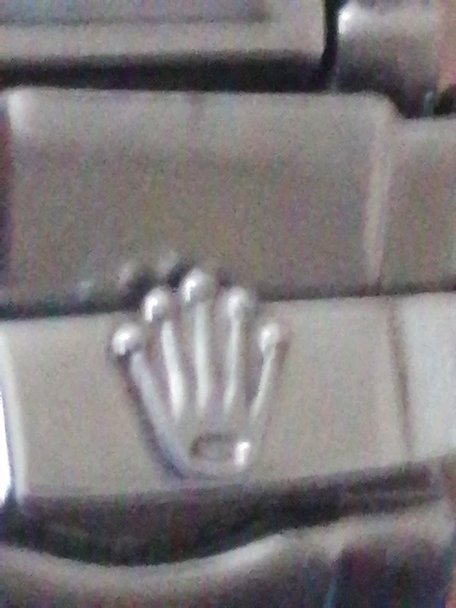The image shows a close-up view of a Rolex crown logo embossed on a metallic surface, likely a watchband or case. The logo consists of five distinct points, representing the five points of a crown. The surface is textured and appears to be brushed metal. The lighting in the image is somewhat diffuse, creating highlights on the raised portions of the crown. There are no people or any other objects visible in the frame, nor is there any context suggesting a location within a city.
