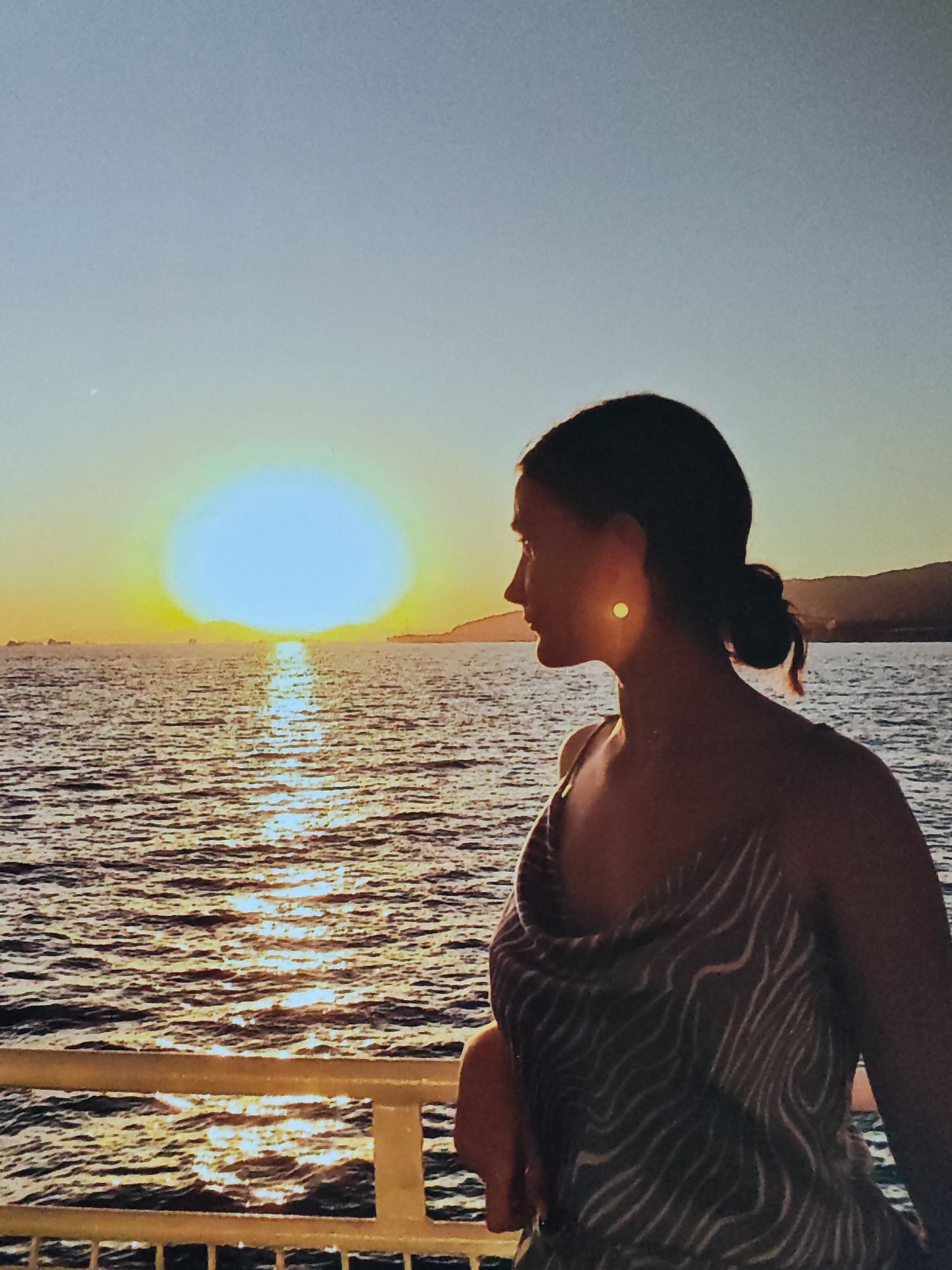 A silhouette of a woman with her hair in a bun is seen from the side, facing the ocean at sunset. She is wearing a patterned dress. The sun is setting over the ocean, casting a reflection on the water. In the background, there are hills on the horizon. The woman is leaning on a railing.