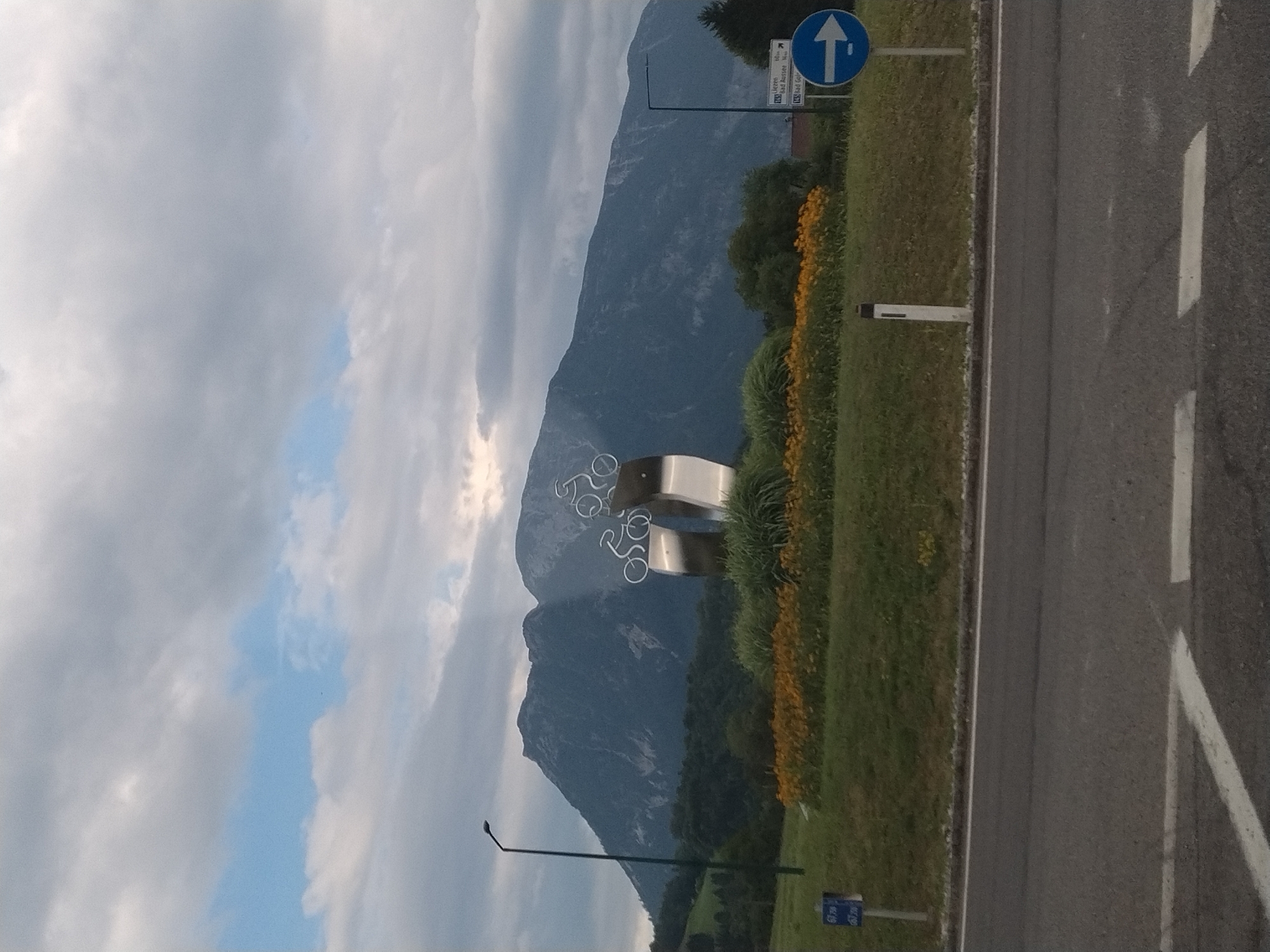 The image depicts an outdoor scene with a large mountain range in the background under a cloudy sky. In the mid-ground, there is a silver sculpture with bicycle motifs and a flower bed with yellow flowers. A road with white markings runs across the foreground. There are traffic signs and a lamppost visible. The location is Gschwandt, Austria, as indicated by the road signs which show directions to Liezen, Bad Aussee, and Bad Goisern.