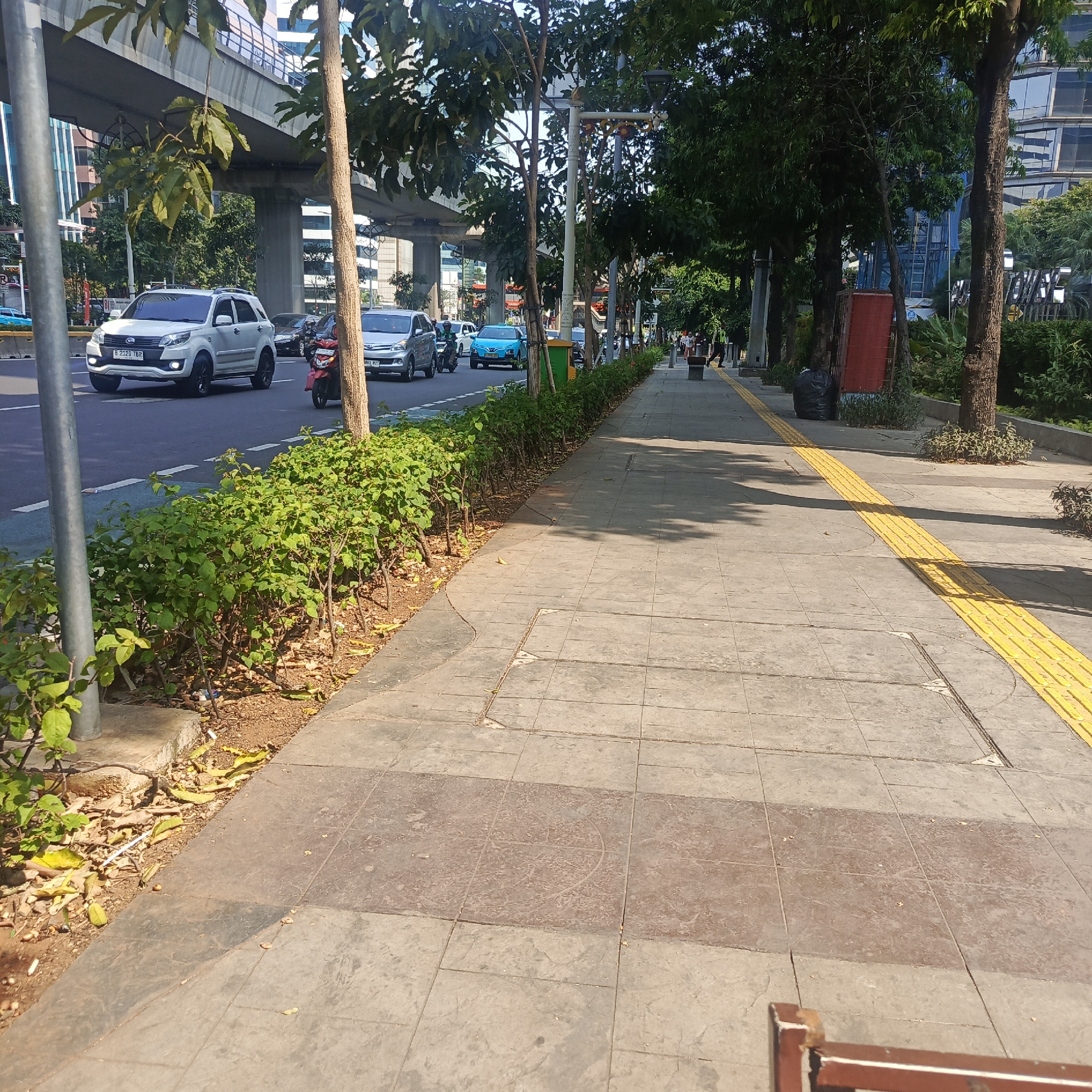 This is a ground-level, eye-level, medium shot of a sidewalk and street scene in South Jakarta, Indonesia, on a sunny day. The sidewalk, paved with large square concrete tiles and featuring a tactile paving strip of raised yellow bumps for the visually impaired, runs along the right side of the frame. Trees line the sidewalk, casting shadows on the pavement. Beyond the sidewalk is a busy street with several cars, including a white SUV and a blue sedan, and a red motorcycle. An elevated highway structure is visible in the background, along with modern buildings. The scene is characterized by urban infrastructure, vehicular traffic, and pedestrian pathways bordered by greenery. There are no people clearly visible walking on the sidewalk. A black garbage bag is placed near the edge of the sidewalk. The general atmosphere suggests a typical day in a bustling urban environment.