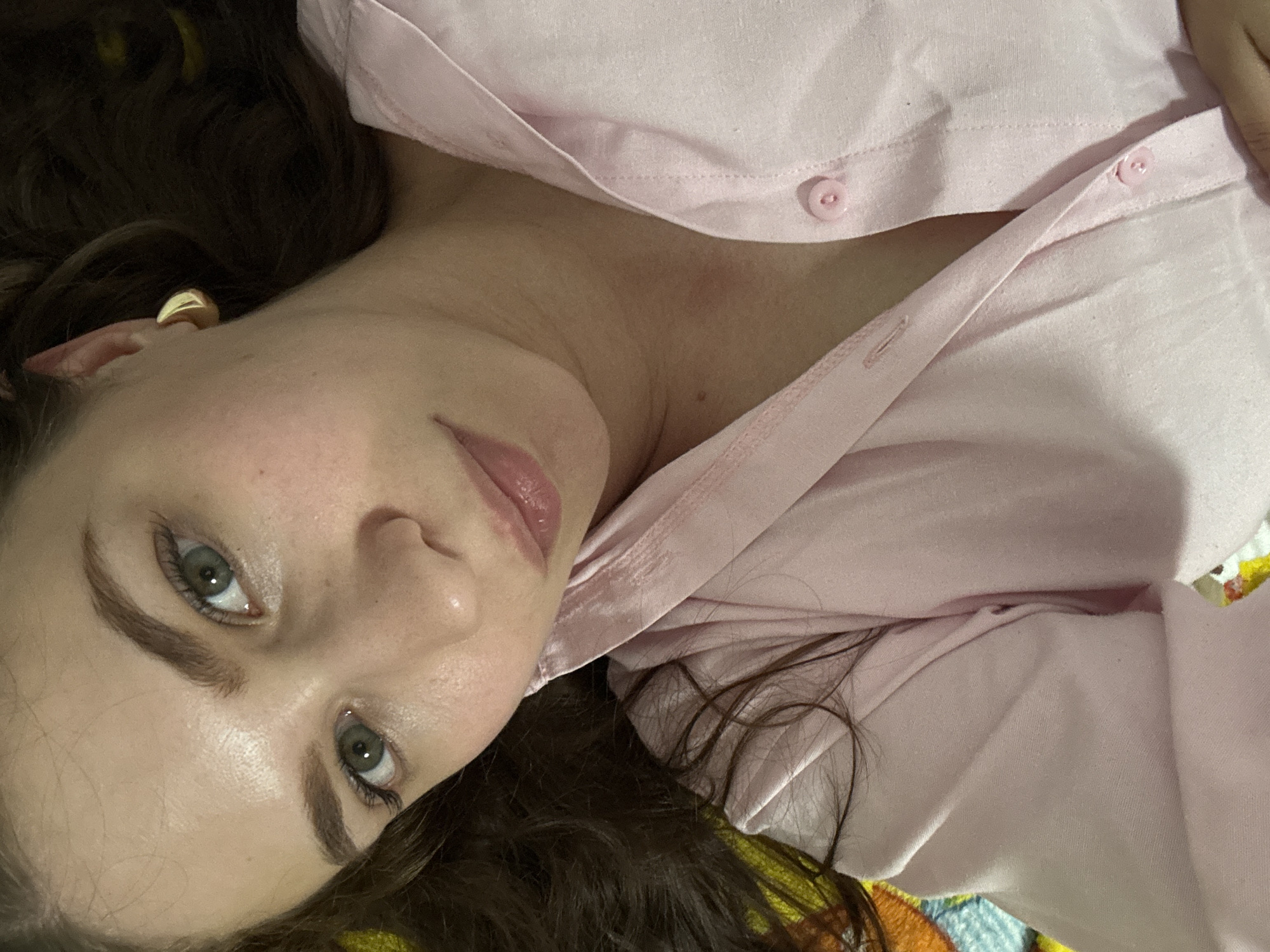A woman lies on her back, looking up at the camera. She has long, dark brown hair that is spread out around her head. She is wearing a light pink button-down shirt. Her eyes are green, and she has a light dusting of freckles across her nose and cheeks. A gold hoop earring is visible in her left ear. The background appears to be a colorful, patterned blanket. The setting is likely a bedroom or living room, indoors. There is no specific information about the location in Chaikovsky, Russia, that can be determined from the image.