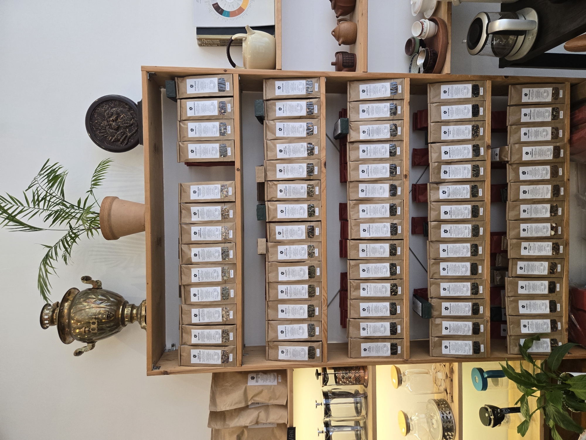 The image depicts a vertically oriented, eye-level shot of a wooden shelf unit against a white wall, seemingly inside a retail setting. The shelves are stocked with numerous small, rectangular packages with labels. Various other objects adorn the shelves above and below.
To the left of the shelving unit, attached to the white wall, is a potted plant with a bronze-colored samovar below it. Above the top shelf are coffee or tea related items and a color wheel chart. Below the bottom shelf are more bagged items and tea brewing equipment.
The overall scene suggests the interior of a specialized tea or coffee shop, possibly in the city of Budva, Montenegro.