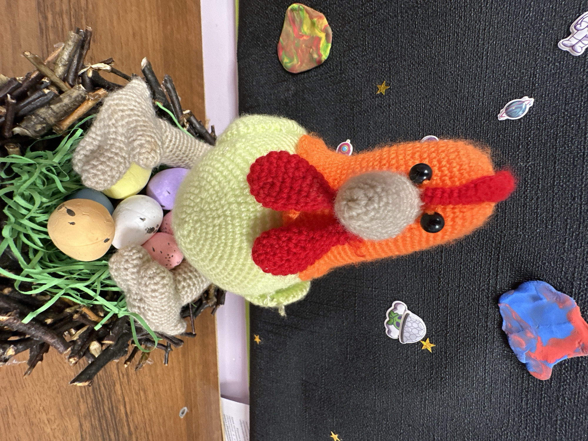 The image depicts a handcrafted Easter scene. On the left, a bird's nest made of twigs and filled with green paper shreds contains several painted Easter eggs in various colors. A crocheted, multi-colored bird (beige wings, light green body, orange head with red tufts, beige beak, and black button eyes) rests partially on the nest. The background is a black surface adorned with space-themed decorations: colorful clay formations resembling planets, stickers of planets and a space suit, and small gold stars. The scene appears to be set indoors. The location in Chaikovsky, Russia is not discernible from the image.