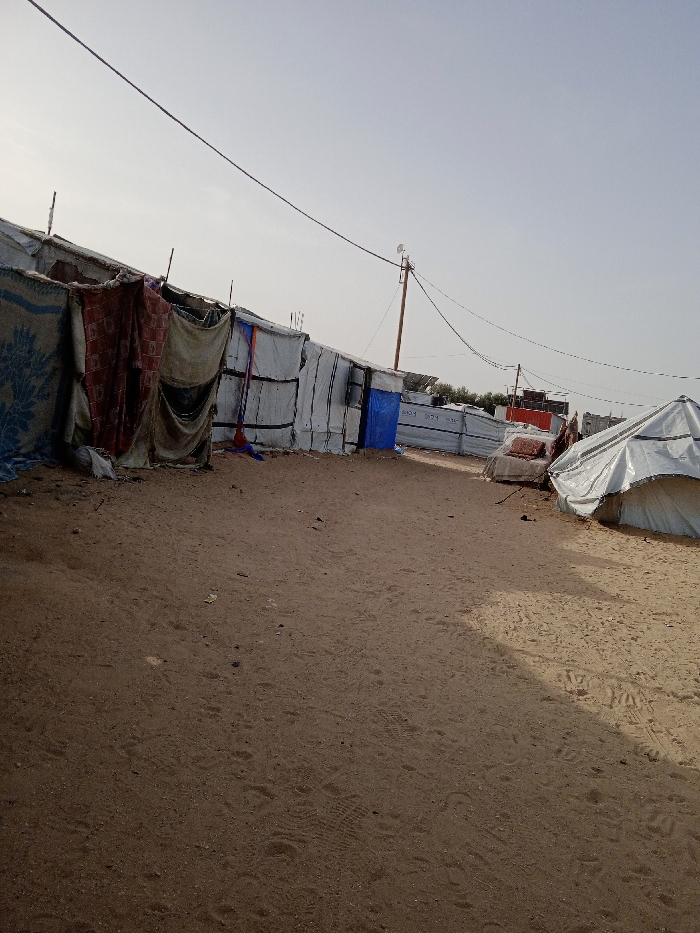 The image displays an outdoor scene in Khan Yunis, Palestinian Territory, characterized by a collection of makeshift shelters and tents constructed on barren, light brown sand or dirt ground under a clear, pale blue sky.
On the left side, a row of improvised shelters extends into the background. These structures are predominantly fashioned from white and grey tarpaulins or plastic sheeting, secured with dark strapping or tape. Visible coverings or door sections include a blue and white patterned blanket, a reddish-brown patterned fabric, and a beige/khaki fabric. One shelter features a prominent blue tarp as a door. Sequential numbers such as "534," "574," and "530-" are discernible on some white tarpaulin panels.
Towards the right foreground, a large white conical or dome-shaped tent, covered in white sheeting, occupies part of the frame. Further behind it, another structure, partially visible, also appears to be composed of similar materials, with a reddish fabric or item on top.
The ground surface is loose sand or dirt, marked by numerous footprints and tire tracks. Small pieces of debris are scattered across the ground. A wooden utility pole stands centrally in the mid-ground, supporting multiple black electrical or communication cables that stretch horizontally across the upper frame. A white, possibly antenna-like, device is mounted at the top of this pole. In the distant background, beyond the shelters, sparse green vegetation and what appear to be solar panels are faintly visible. No individuals are present in the scene.