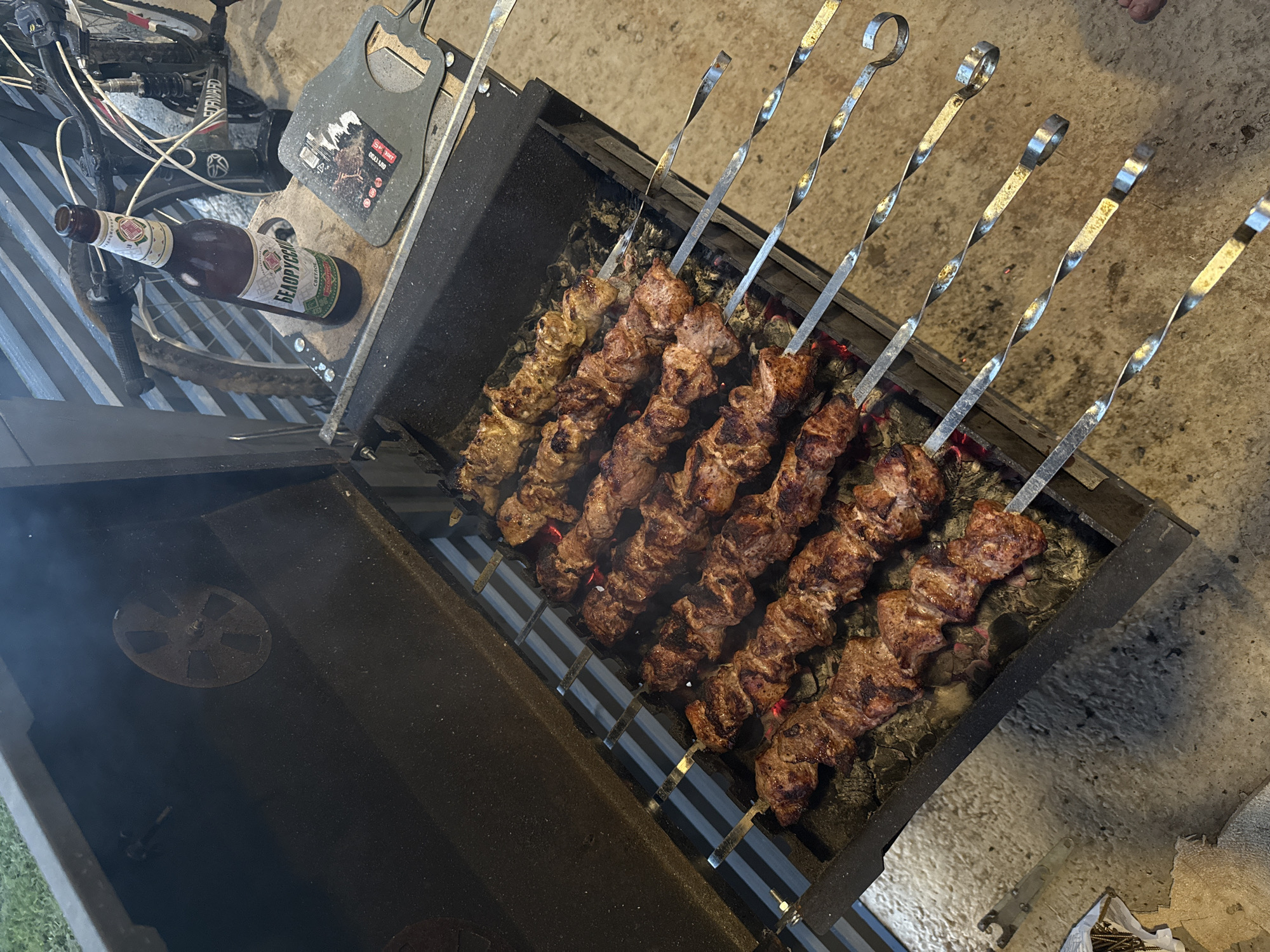 In the city of Podshivalovo, Russia, a barbecue grill is open, with numerous skewers of meat cooking over hot coals. A bottle of Belarusian beer and a bottle opener are placed on the side of the grill. In the background, a bicycle is leaning against a fence. The scene appears to be an outdoor gathering, possibly in a backyard or park, given the presence of a grill and bicycle. The lighting suggests it is either late afternoon or early evening.