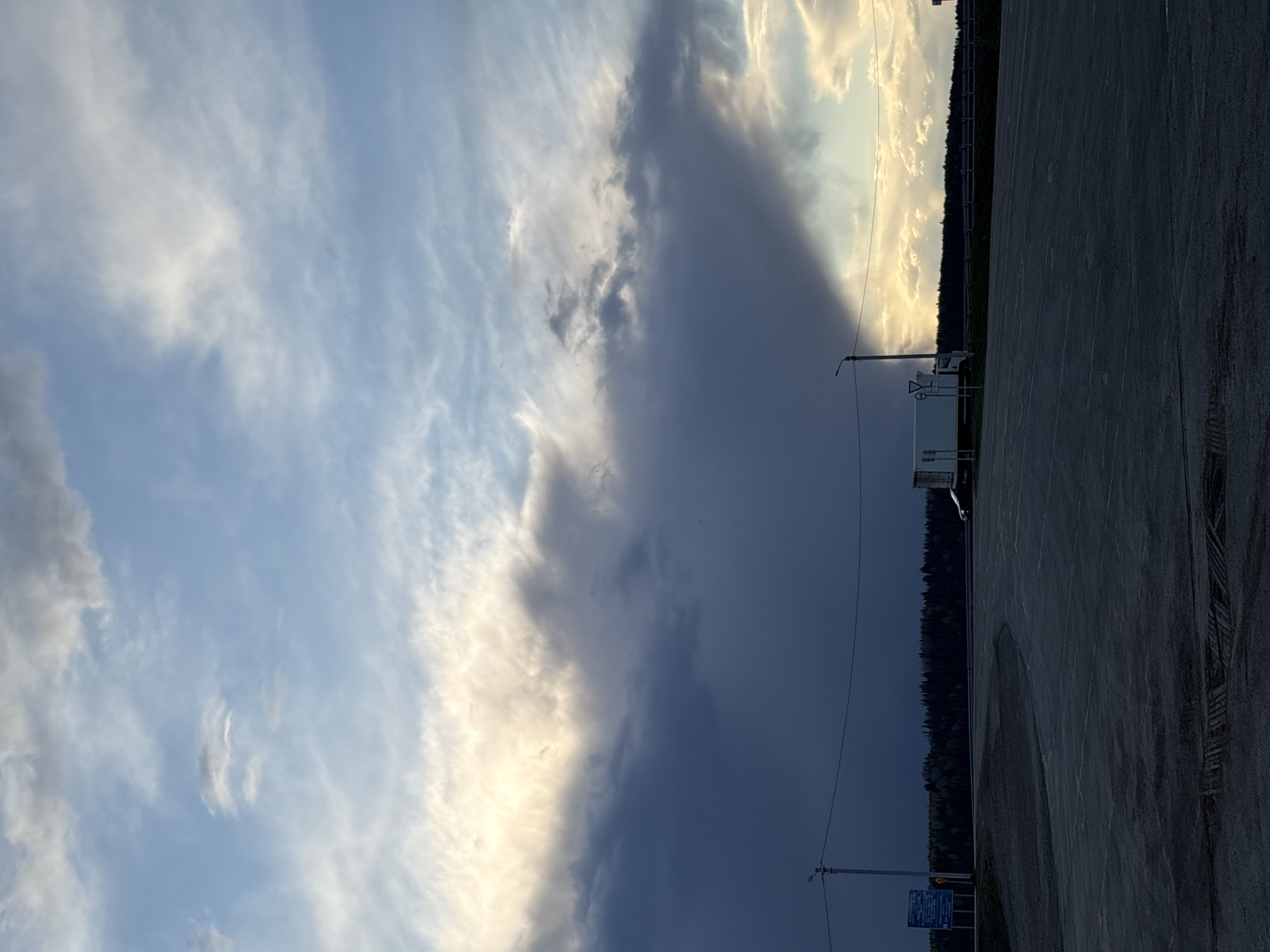 The image presents a view in Chernovskoe, Russia. The sky dominates the scene, displaying a stark contrast between a patch of blue sky with scattered white clouds and an approaching dark, ominous storm cloud. A section of pavement is visible in the foreground. In the middle ground, there's a narrow strip of dense, dark green forest. An electric pole and some road signs stand near a white van. The lighting is uneven, with the part of the sky unaffected by the storm illuminated, casting shadows onto the landscape.