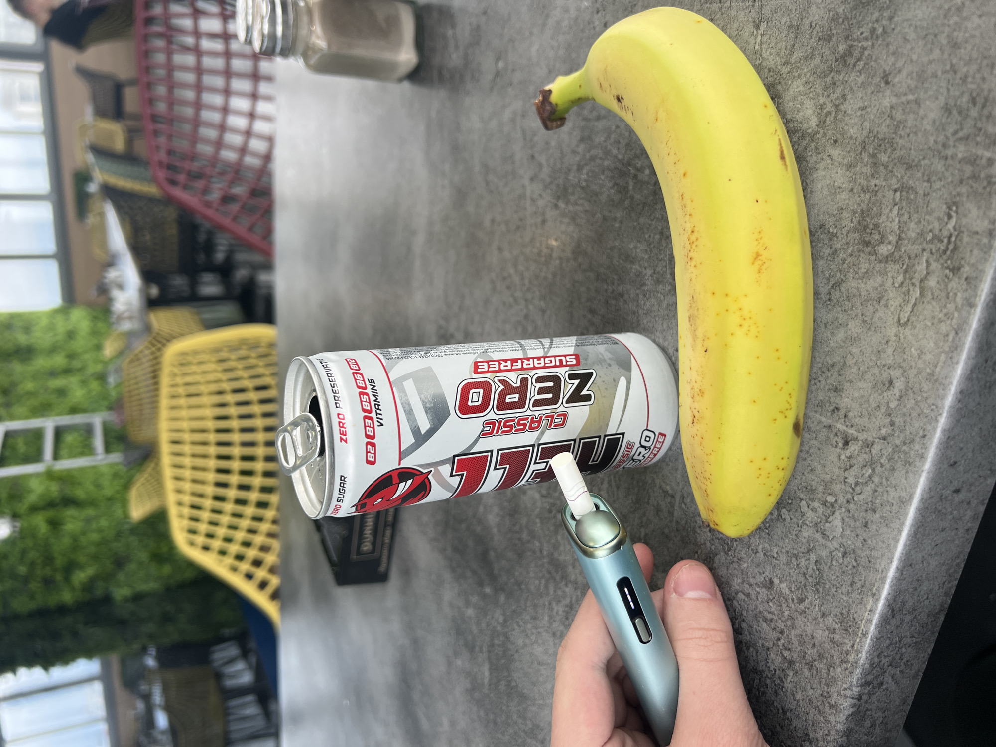 A photograph depicts a still life of items placed on a gray surface. The image appears to have been taken outdoors, possibly on a patio, with a blurred background showing greenery, yellow and red wicker chairs, and a building with large windows. A banana, a can of "Duhill Classic Zero" energy drink, and a blue vape are the primary subjects. A hand is holding the vape. The image suggests a casual setting. There is no information in the image to suggest it was taken in Sofia, Bulgaria.