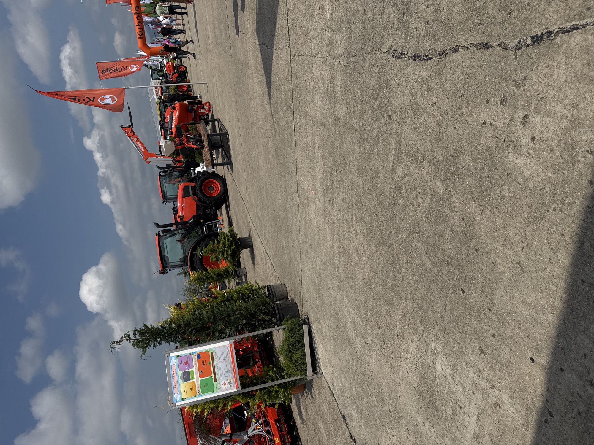 The image displays an outdoor exhibition of agricultural machinery, likely a fair or demonstration, featuring numerous Kubota brand tractors and related equipment. The scene is set on a large, grey paved area, possibly an exhibition ground, under a bright, partly cloudy sky.
In the foreground and extending into the midground, several bright orange Kubota tractors of various sizes are neatly arranged. Some appear to have attachments, such as a front loader or telehandler. Prominently displayed throughout the area are numerous orange flags and banners bearing the "Kubota" logo, with one large triangular flag standing tall towards the left of the frame. In the background, an inflatable archway, also orange and branded "Kubota," is visible.
Several people are scattered in the background, observing the machinery and interacting at the event. Potted evergreen trees and shrubs are placed intermittently among the exhibits, adding greenery to the setting. A significant white sign, standing on a metal frame, is visible in the foreground, showcasing colorful graphical elements and the clear text "KUBOTA PARK" at its top.
The time of day appears to be daytime, suggested by the bright illumination and visible shadows. The weather is fair, with a blue sky dotted with white cumulus clouds. The overall atmosphere suggests a relaxed, informative event focused on agricultural equipment. The event takes place in Stara Zagora, Bulgaria.