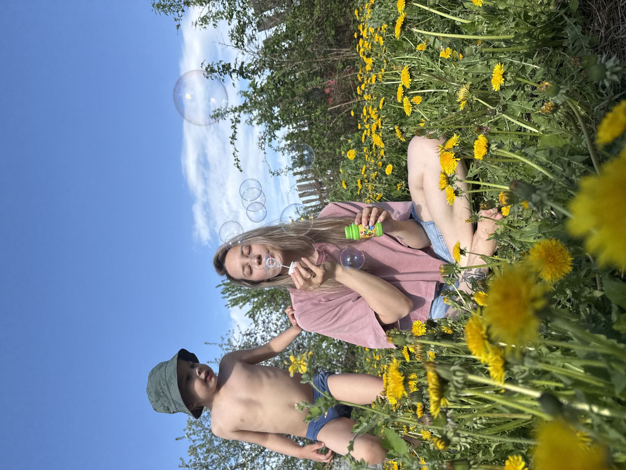 A woman and a young child are in a field of dandelions on a sunny day. The woman is sitting in the field, blowing bubbles. She is wearing a pink t-shirt and denim shorts. The child is standing next to her, reaching for the bubbles. He is wearing a green hat and blue swim trunks. The sky is blue with a few clouds. The location is Chaikovsky, Russia.