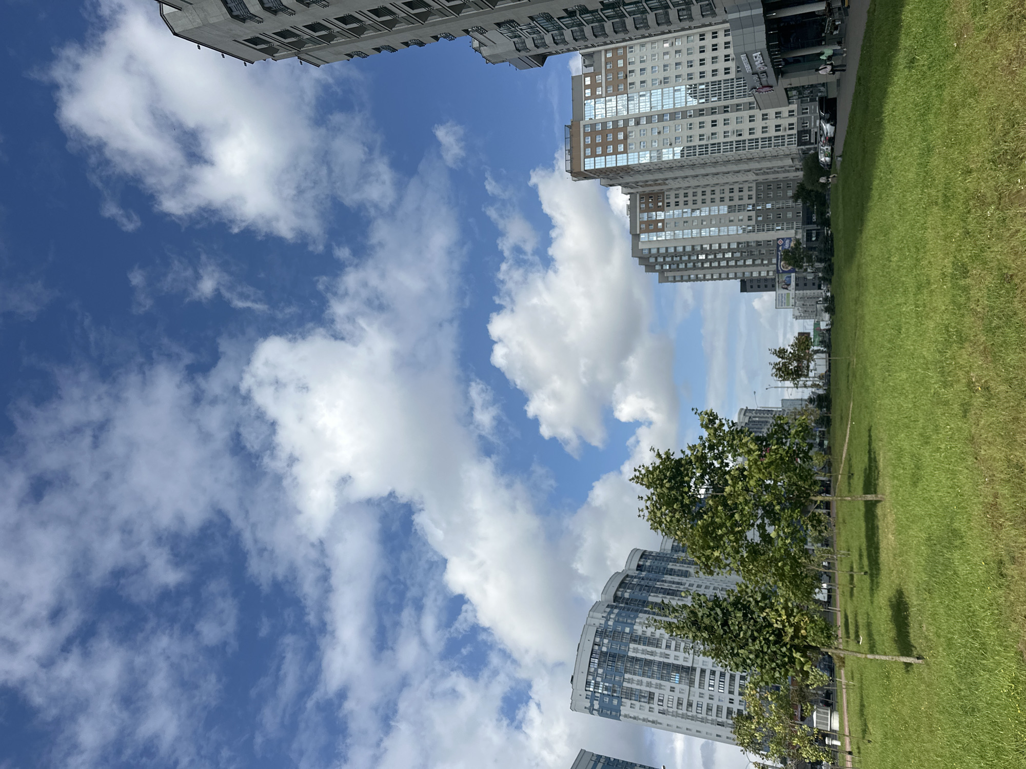 This is a medium shot, eye-level, outdoor, daytime, urban landscape photograph. The scene depicts a modern residential area with several high-rise buildings under a bright blue sky with scattered white clouds. In the foreground, a neatly mown, bright green lawn is dotted with young trees that cast shadows. Beyond the lawn, a row of modern apartment buildings with numerous windows is visible. The architecture is characterized by a mix of grey and blue facades, with some buildings featuring curved designs. A street with parked cars and a few pedestrians is visible in the midground, leading towards the buildings. The overall atmosphere is one of a contemporary urban environment on a clear day, likely in Minsk, Belarus, given the architectural style and the presence of Cyrillic script on signage.