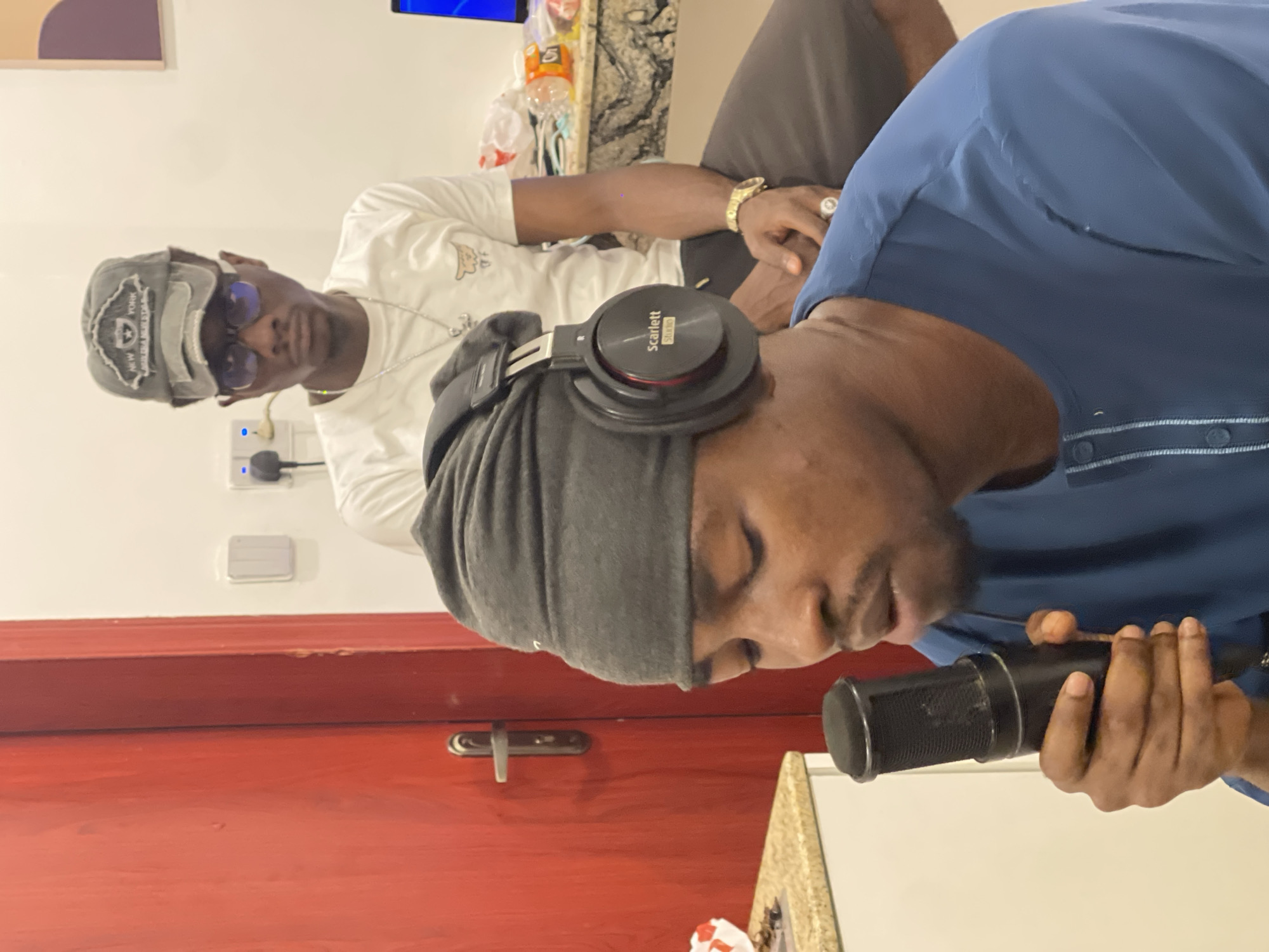 Two individuals are present in what appears to be an indoor setting, possibly a studio or recording space, given the presence of audio equipment. The primary subject in the foreground is wearing a blue collared shirt and headphones over a grey beanie. They are holding a black microphone with both hands, suggesting they might be speaking into it or adjusting it for recording.
The second individual, situated behind and to the left of the foreground subject, is wearing a white t-shirt and a grey baseball cap. They are also wearing sunglasses. This individual appears to be looking towards the camera.
Notable objects include a pair of black Scarlett Studio headphones. The background shows a white wall with some electrical outlets and a red wooden door with a silver handle. There is also a glimpse of a marble-patterned countertop.
The lighting suggests it is daytime, and the overall atmosphere seems casual, possibly related to a podcast recording, music production, or a similar audio-focused activity. No specific weather or time of day indicators are visible beyond the interior lighting. The facial expressions of both individuals are somewhat obscured or neutral.
Visible text includes "Scarlett Studio" on the headphones. The baseball cap has text and a logo that are partially legible, but appear to read "NEW YORK".