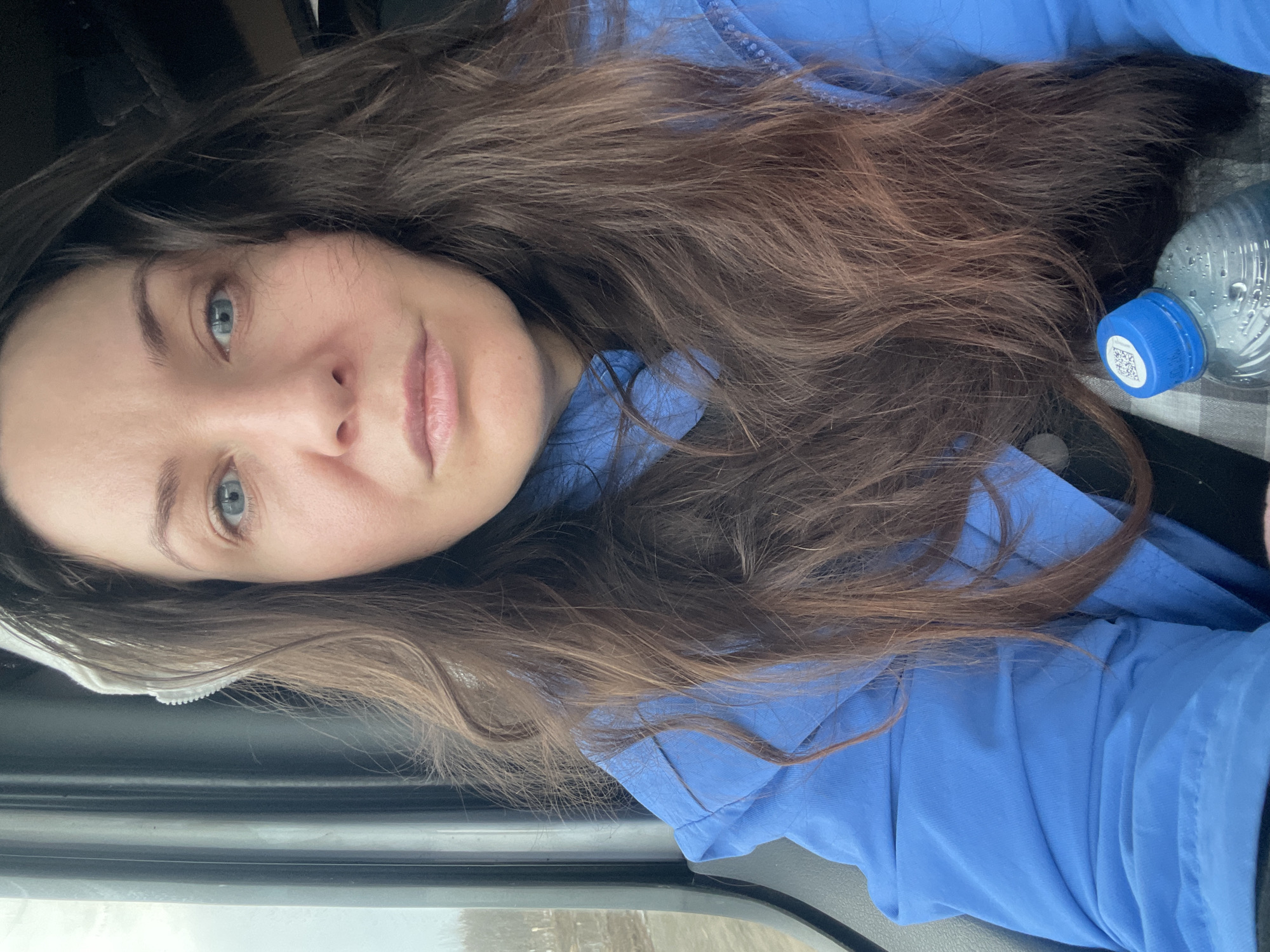 A woman with long brown hair and blue eyes is pictured indoors, wearing a blue shirt. A water bottle with a blue cap is visible to her right. The scene is possibly inside a vehicle due to the presence of a car window frame. There is no information in the image to determine location in Yus'ki, Russia.