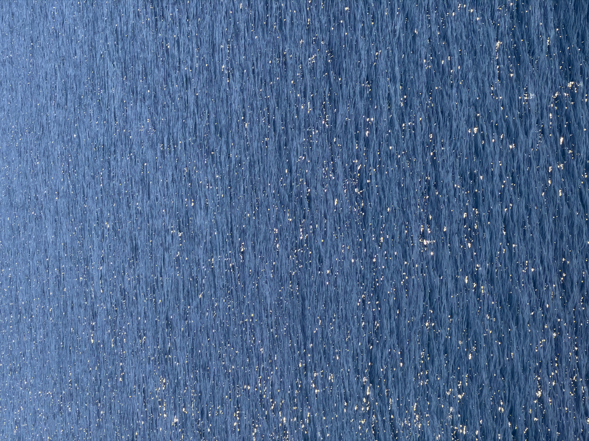 This image captures a close-up view of the Adriatic Sea near Petrovac na Moru, Montenegro. The water is a deep blue, its surface broken by countless small waves. Sunlight reflects off the crests of these waves, creating scattered glimmers of light that appear like tiny stars across the water's surface. The texture of the water is rough and slightly uneven due to the wind-driven wavelets. There are no discernible people, objects, or specific subjects beyond the seascape itself. The scene's focus is entirely on the interplay of light and water, conveying a sense of movement and natural beauty.