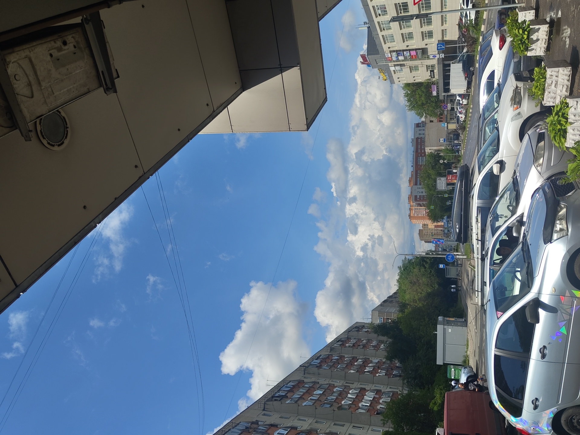 The image depicts a street scene in Zhukovsky, Russia, on a partly cloudy day. In the foreground, a silver car with holographic geometric decals on its side is parked, along with other vehicles in a parking lot. Behind the cars, a multi-story apartment building with numerous windows is visible on the left. Trees and other buildings are scattered throughout the mid-ground. The sky is a vibrant blue with scattered white clouds. The scene suggests a typical urban environment.