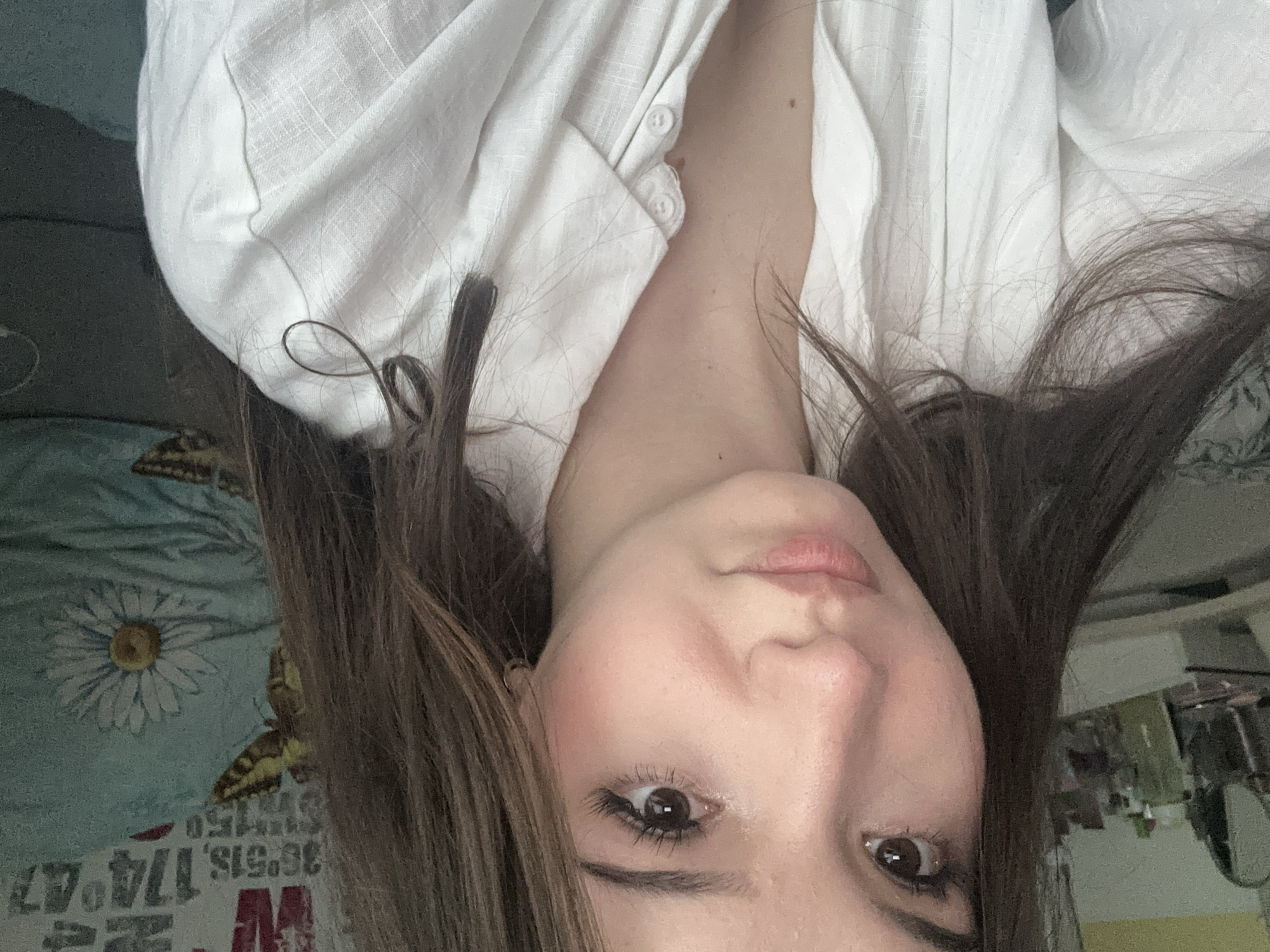 A young woman with long, brown hair is pictured from the chest up, looking directly at the camera. She is wearing a white button-down shirt. Her hair is slightly messy, with some strands falling forward over her shoulders. She is wearing subtle makeup, including mascara and blush. Behind her, a blue pillow with a white daisy pattern is visible. The lighting in the image is soft and natural, suggesting an indoor setting. There is no discernible information within the image that would indicate a specific location in Naberezhnye Chelny, Russia.