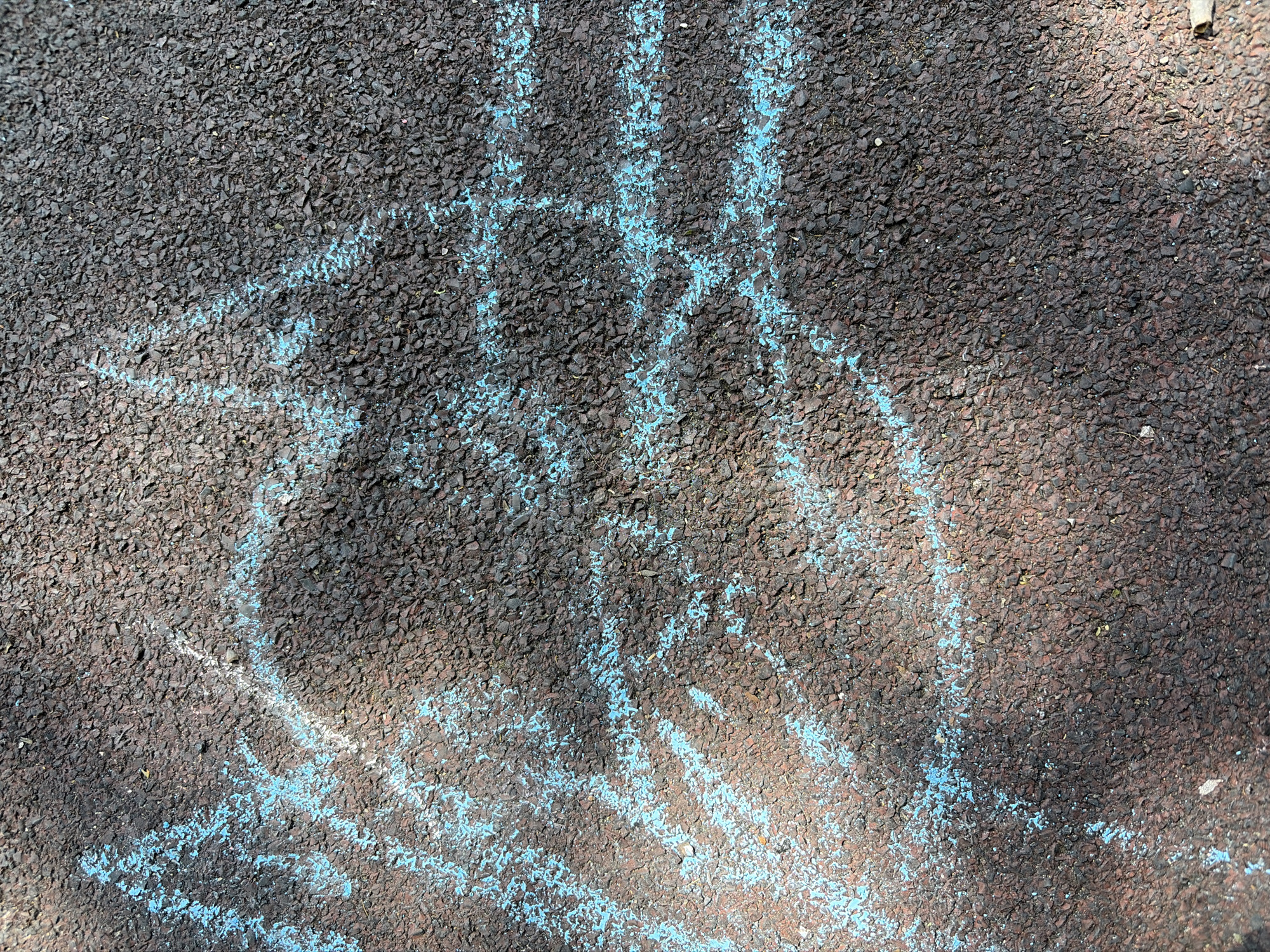 A top-down shot of dark-colored asphalt in Svetovrachene, Bulgaria, shows a faded drawing in blue chalk. The drawing appears to be a crude hopscotch grid with irregular lines and shapes. The asphalt texture is rough, and the chalk lines are broken and uneven, indicating wear. There are shadows on the right side of the image, suggesting the direction of the light source.