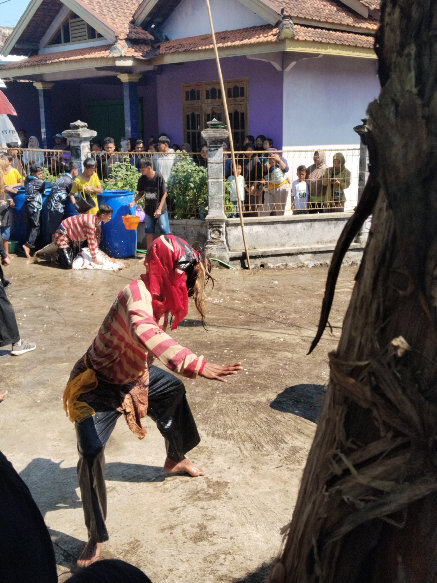 In an unidentified urban area of Unknown, Unknown, a man with a red cloth over his head, dressed in a striped long-sleeve shirt, dark pants, and a yellow sash, stands barefoot on a wet surface. His arms are extended, palms down, as if performing or reacting. Behind him, another man, also in a striped shirt, is bending over, seemingly involved with a white animal (possibly a goat). Several individuals, including men and women, some with headscarves, are gathered behind a low wall, observing the scene. Blue barrels are scattered near the wall, with people holding buckets. The environment includes a two-story building painted in lavender and white and a large tree trunk in the foreground, slightly obscuring the view. The ground is wet and speckled, suggesting a recent or ongoing water-related activity.