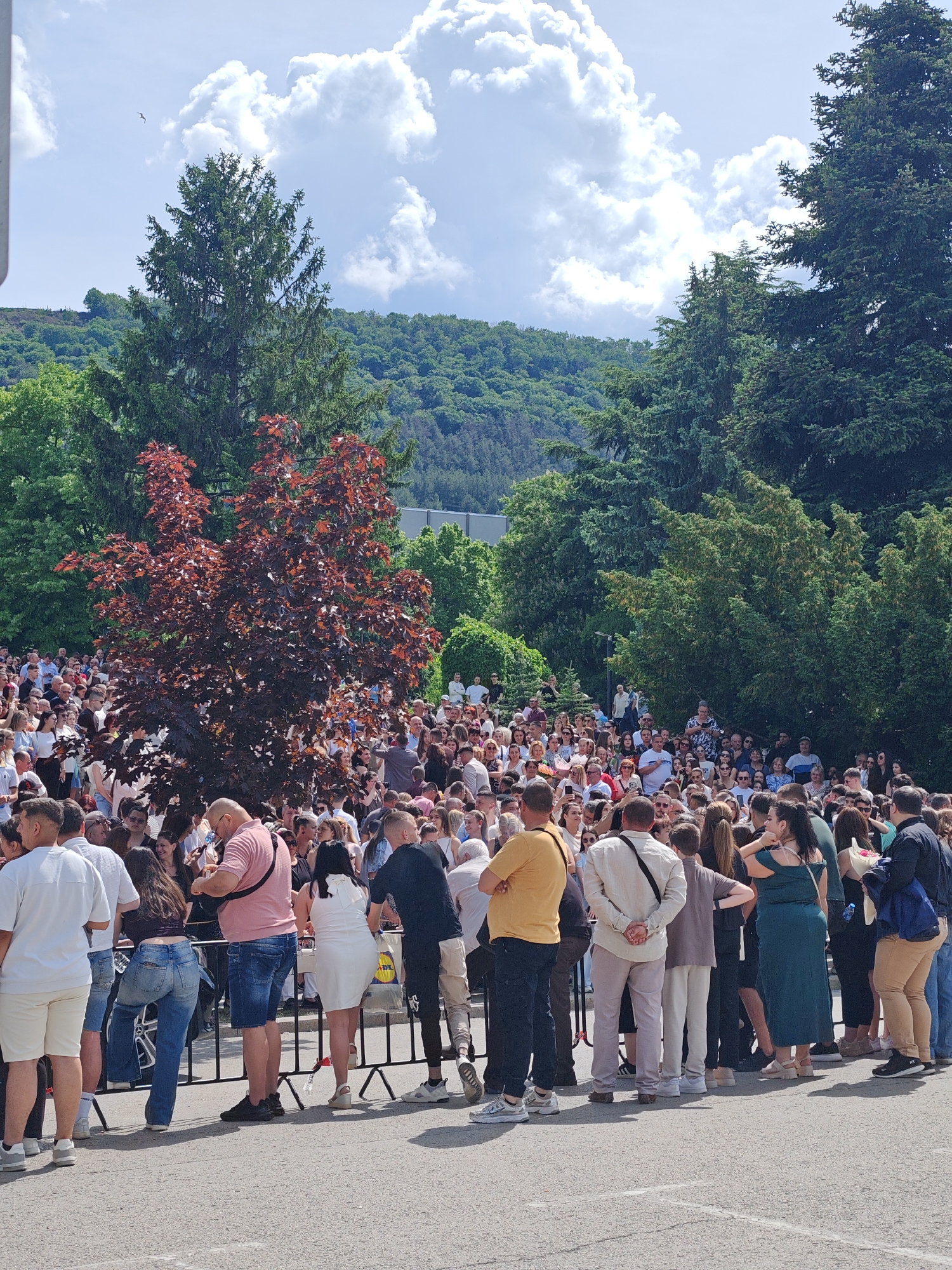 The image captures a large crowd of people gathered outdoors in Shumen, Bulgaria. A metal barrier separates the crowd from the foreground. Individuals of various ages and attire are visible, with many appearing to be standing and waiting. The background features lush green trees and foliage, along with a partial view of hills in the distance. The sky is partly cloudy.