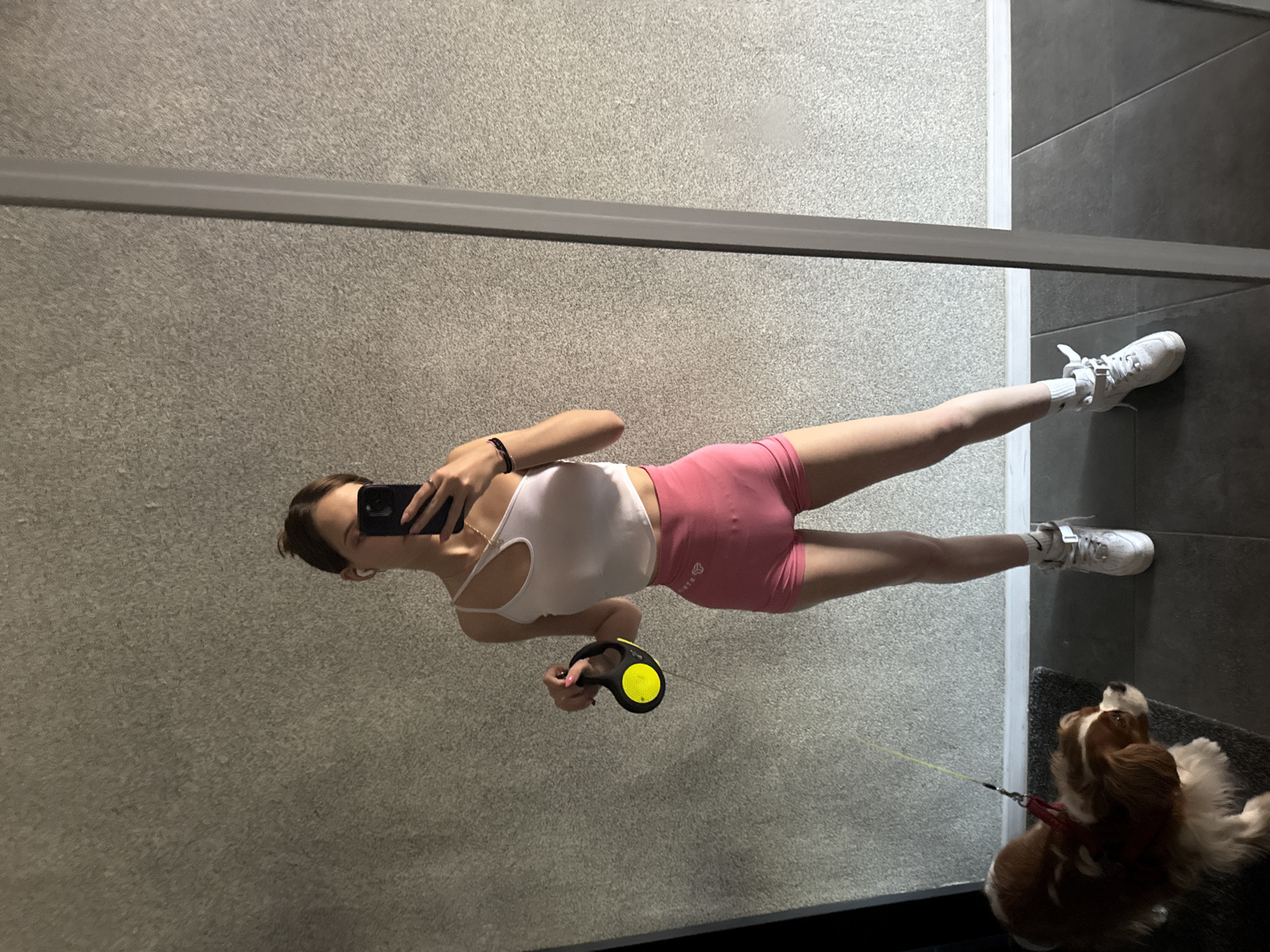 A young woman, seen in a full-body mirror reflection, stands in what appears to be a hallway or entryway. She is wearing a white asymmetric crop top, pink high-waisted shorts, and white high-top sneakers. She holds a black retractable dog leash with a yellow circular detail in her left hand. A Cavalier King Charles Spaniel, wearing a red harness, is visible in the lower left corner of the frame, on a dark grey doormat. The background consists of a textured grey wall and dark grey floor tiles. A vertical grey column, likely part of a doorframe or mirror frame, is present on the right side of the reflection. The lighting suggests an indoor setting, possibly in a residential building or gym, in Sofia, Bulgaria.