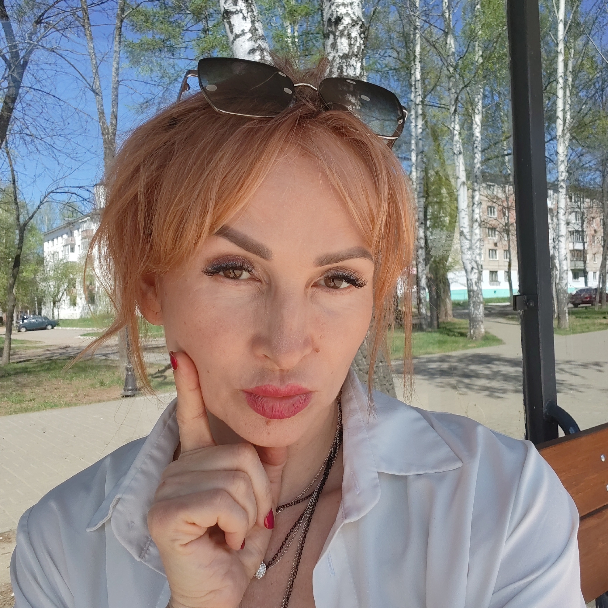A woman is seated on a park bench in Chaikovsky, Russia. She is fair-skinned with reddish-blonde hair and sunglasses perched on her head. She wears a white button-down shirt and a layered necklace. Her left hand is raised to her chin, with her index finger extended upwards and touching her cheek. Behind her, a park scene with birch trees, grass, and a paved walkway unfolds. In the background, a multi-story apartment building is visible, along with parked cars. The weather appears sunny and clear.