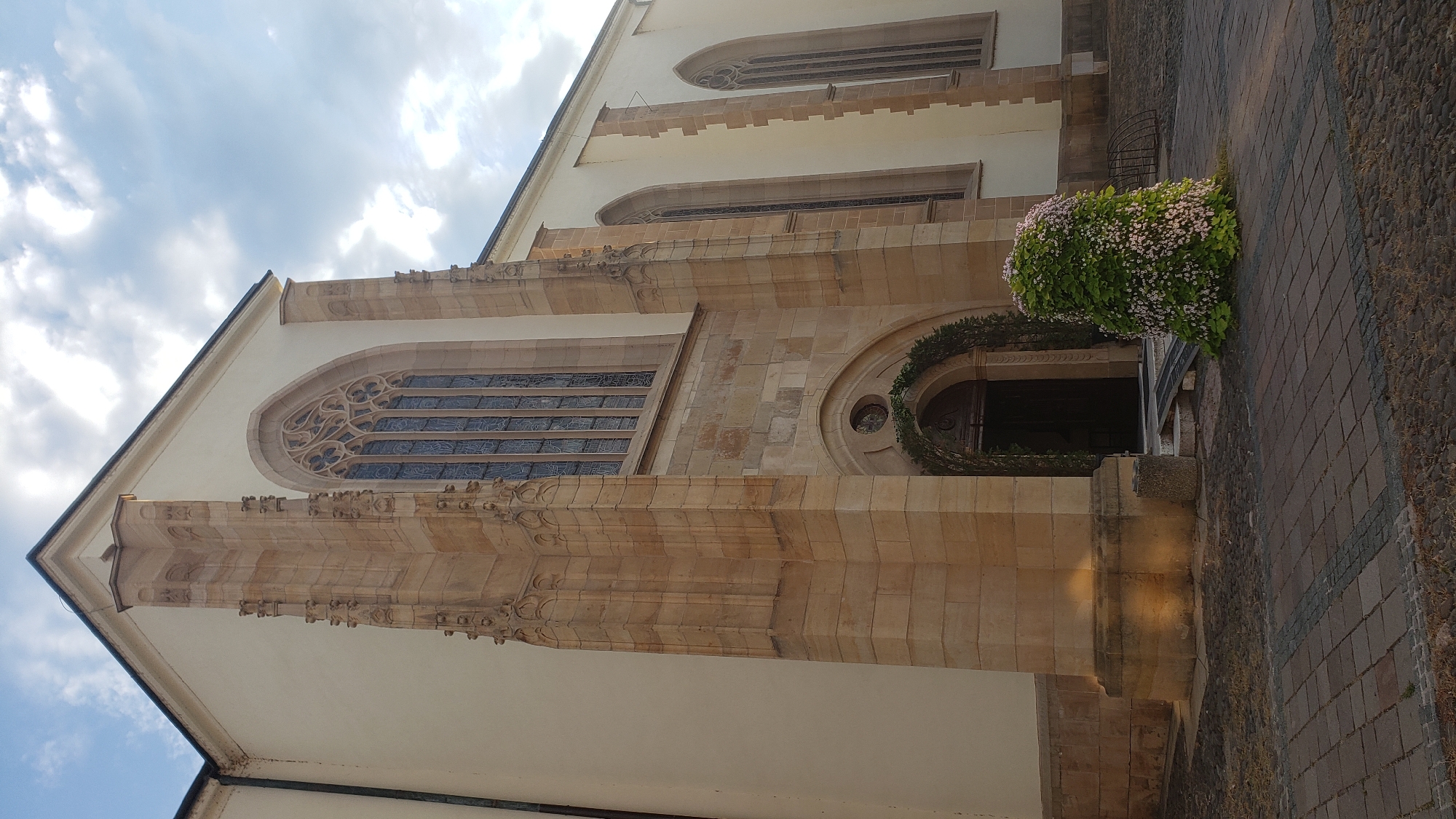 The image shows the exterior of a large, light-colored building with Gothic architectural elements. The building is constructed of stone and has tall, arched windows with stained glass. A prominent entrance features an arched doorway decorated with a green wreath. To the right of the entrance, a large, bushy plant with small pink flowers is positioned. The ground in front of the building is paved with cobblestones and brickwork. The sky above is partly cloudy with sunlight filtering through. Based on the architectural style and surrounding environment, this building is likely St. Nicholas Cathedral in Prešov, Slovakia. There are no people visible in the image.
