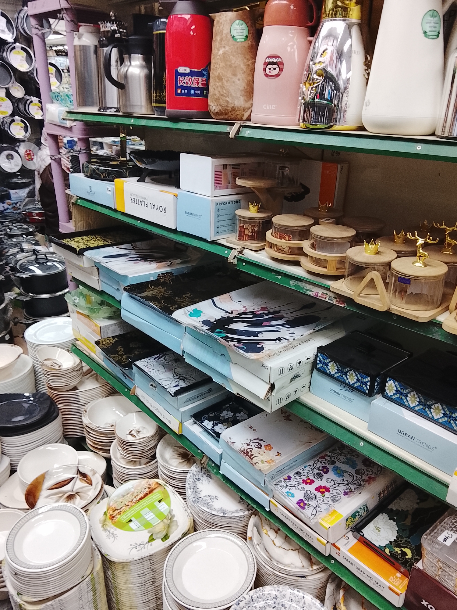 The image captures a retail scene in Gulgasht Colony, Pakistan, featuring shelves stacked with dishware, containers, and serving items. The shelves are organized with metal structures painted in a light purple hue, and the shelf surfaces are green. The top shelf displays thermoses and containers, with brands such as "Cille" visible. Below, boxes labeled "ROYAL PLATTER" and "URBAN TRENDS" are arranged. Several wooden-lidded containers are positioned as well, some with decorative gold accents.
On the lower shelves, stacks of plates and bowls, featuring various designs and patterns, dominate. Plates in white, gray, and decorated styles are neatly piled, while boxes labeled "SMART SERVING TRAY" and others with the "URBAN TRENDS" brand are also present. The environment suggests a busy retail space specializing in tableware and kitchenware.