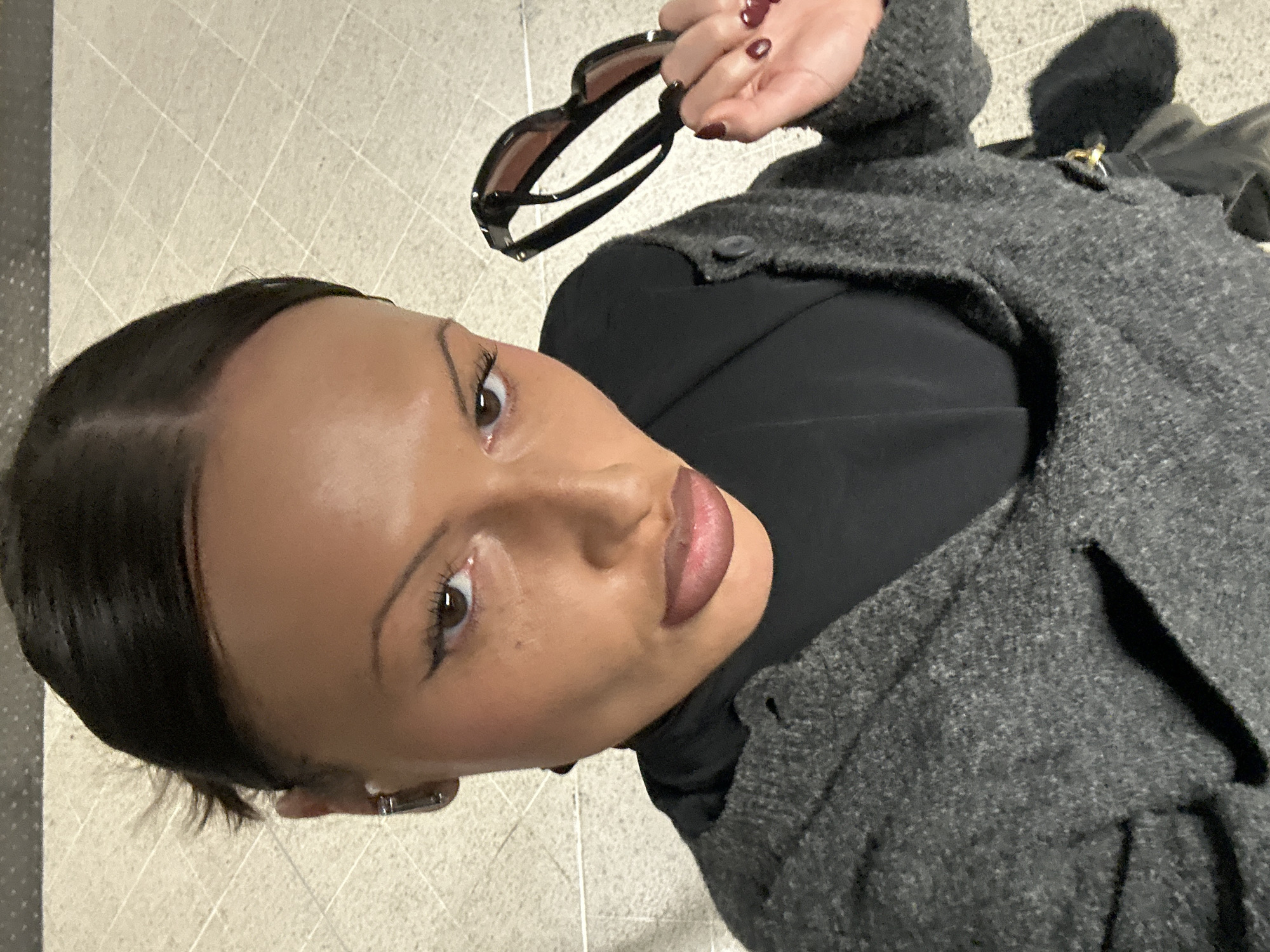 A woman with dark hair pulled back, wearing a black turtleneck and a grey wool coat, holds sunglasses in her hand. The photo is taken from above, looking down at the woman. She appears to be standing on a tiled floor. The location is indeterminable.