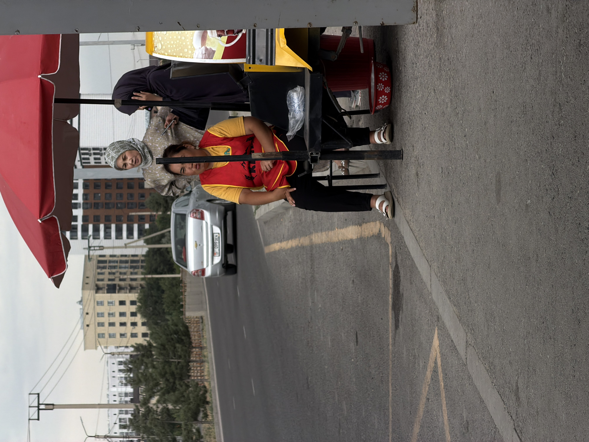 The image shows three people standing near a street vendor's cart. The cart has a red umbrella and a yellow awning. One person, wearing a dark headscarf and a light-colored patterned robe, is leaning on the cart. Another person, wearing a yellow shirt and a red vest, is also standing by the cart, looking towards the camera. A third person, wearing a hijab and a light-colored patterned robe, is standing to the left of the cart, also looking towards the camera. A silver car is parked on the street behind the cart. The scene appears to be outdoors, with buildings and trees visible in the background. The sky is overcast, suggesting a cloudy day. The people appear to be interacting with each other or waiting for customers. There is no visible text in the image.