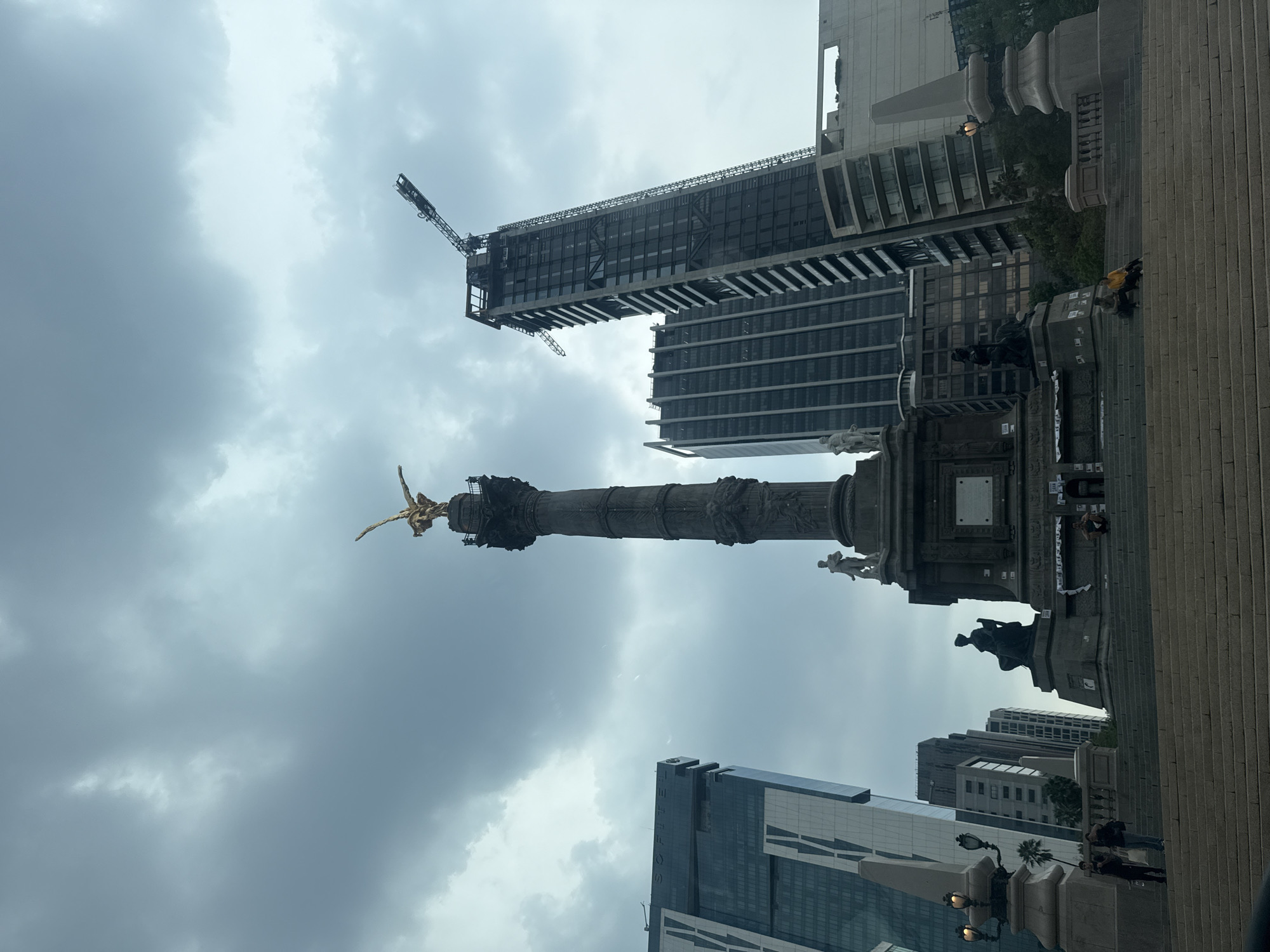 The image shows the Angel of Independence monument in Mexico City, Mexico, against a cloudy sky. The monument is a tall column topped with a golden statue of a winged figure. Several modern buildings surround it. A construction crane is visible next to one building. Several people are walking on the steps of the monument. Additional people are visible closer to the base of the monument and in the vicinity of a street light.