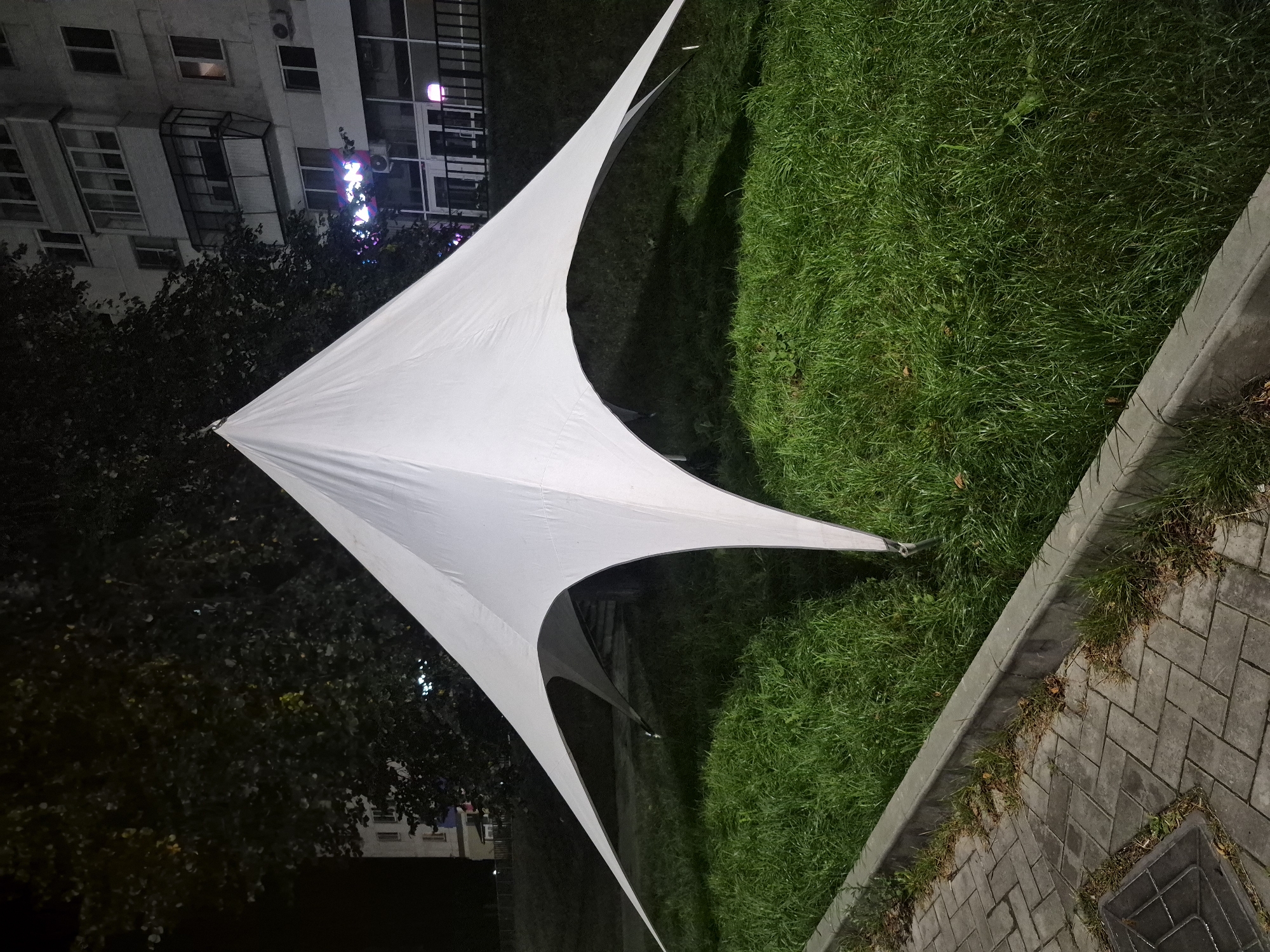 A white, four-pointed, tensile fabric structure, possibly a tent or canopy, is erected on a grassy area at night. The structure has a central peak and four elongated points extending outwards, with the fabric draped in a flowing manner. The ground beneath the structure is covered in green grass. In the foreground, a paved walkway made of gray rectangular blocks runs along the edge of the grassy area, separated by a concrete curb. Beyond the structure and grass, a multi-story apartment building is visible, with lit windows and a neon sign that appears to read "AKON" in purple and pink letters. Trees with dark foliage are present behind the structure and to the left, obscuring parts of the building and the night sky. The scene appears to be an urban setting at night, likely a public space or park area in Chaikovsky, Russia, given the context. There are no people visible in the image.