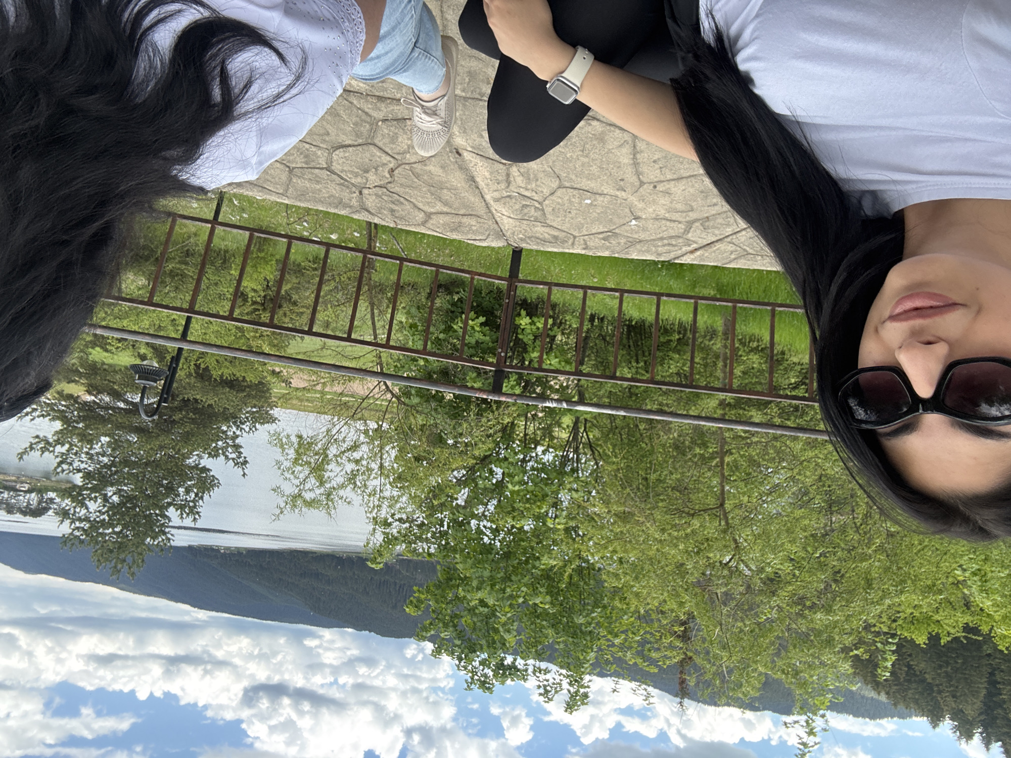 A topsy-turvy eye-level photograph depicts two individuals overlooking a scenic landscape in Dospat, Bulgaria. One person is on the right edge, seen from the nose up, wearing sunglasses, a white t-shirt, and having long dark hair. To the left is another person partially visible; they are wearing a white blouse and light blue jeans with light-colored sneakers. Both are sitting on a stone surface with a railing.
Beyond the railing are trees, a lake, mountains, and a cloudy sky. The composition is askew, giving the viewer the impression of a tilted perspective.