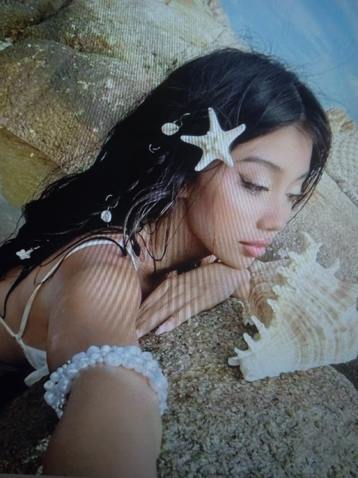 The image shows a young woman with long, dark hair adorned with a starfish hair clip and small dangling charms. She is lying on a sandy surface next to a large seashell. The woman is wearing a white top with thin straps and a bracelet made of white beads on her wrist. Her eyes are closed, and her head is resting on her hand. The background appears to be rocks and a blurred blue sky. There is no discernible location in Chernivtsi, Ukraine.