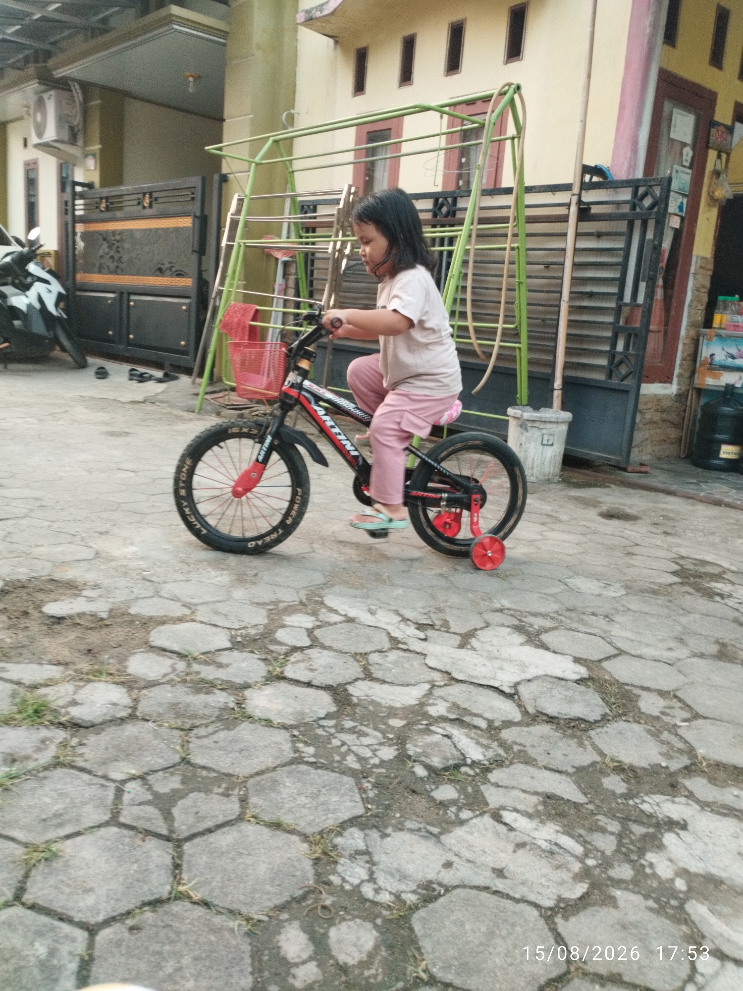 A young girl rides a small bicycle on a paved area outside a building. The girl is wearing a light-colored t-shirt and pink pants. The bicycle is red and black with a pink basket and training wheels. The paved area is made of hexagonal concrete tiles with some grass growing in the cracks. In the background, there is a house with a yellow facade and a green metal structure that appears to be a play area or drying rack. A black motorcycle is parked to the left of the frame, and a black metal gate is also visible. The scene suggests a residential setting in Bandar Lampung, Indonesia. The timestamp on the image reads "15/08/2026 17:53".