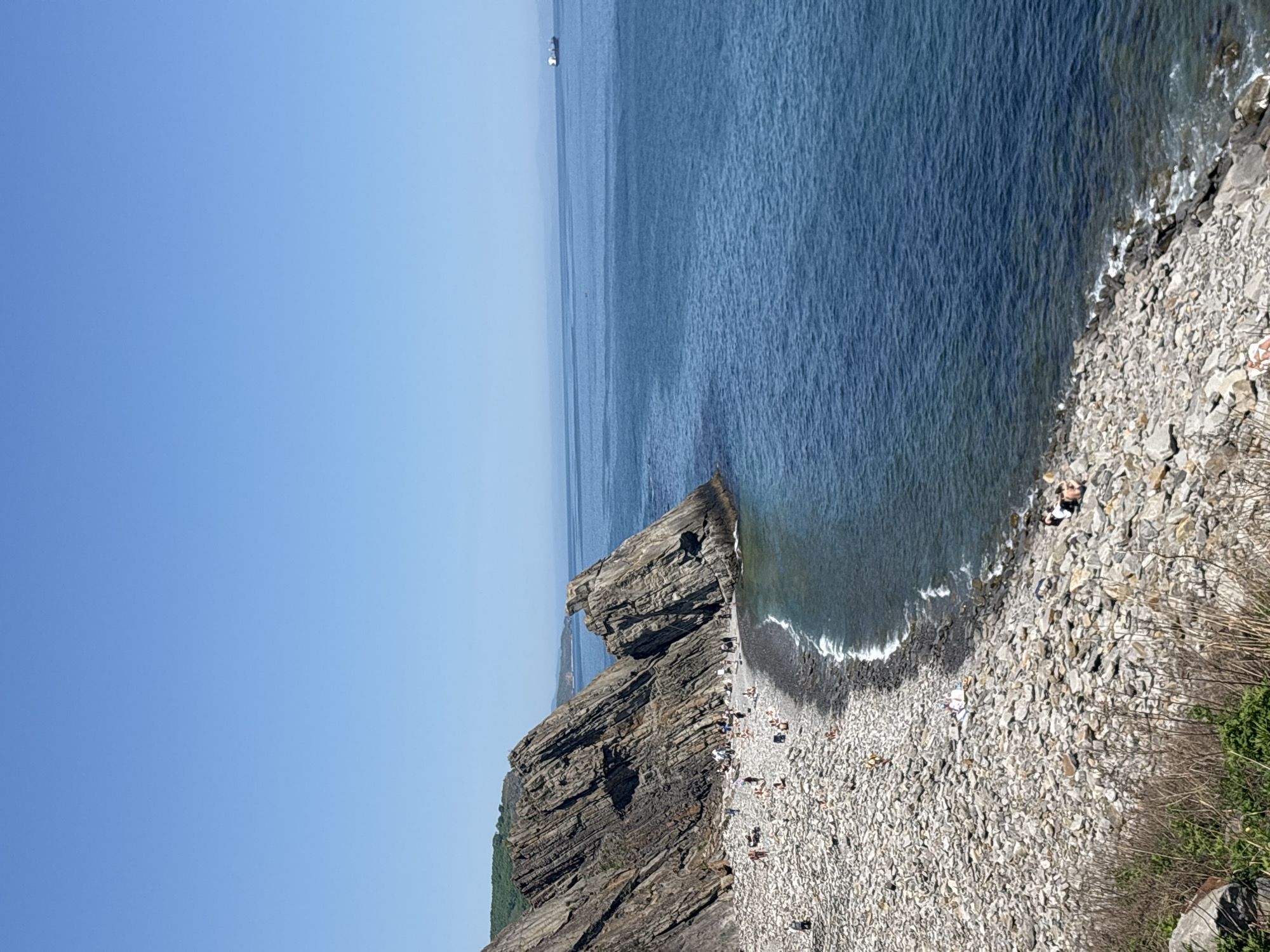 The image depicts a rocky beach in Vladivostok, Russia. A large rock formation divides the beach from the open ocean. People are spread across the pebble-covered beach, some sunbathing and others near the water's edge. The sea is a dark blue with a ship visible in the distance. The sky is clear and light blue. Green foliage lines the edge of the beach on the right.