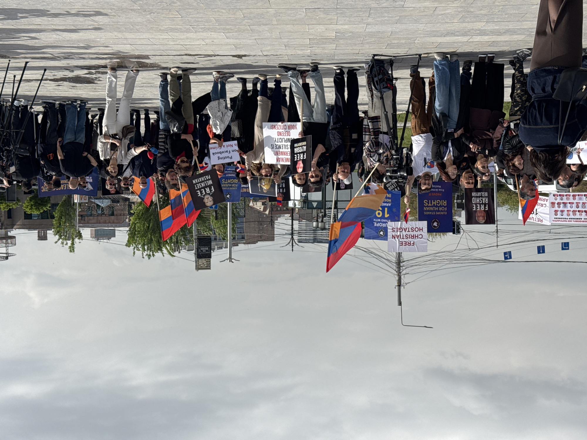 A demonstration is taking place in Yerevan, Armenia, on a paved plaza in front of "The Bridge" building. Approximately 25 people stand shoulder-to-shoulder holding signs and Armenian flags. Signs indicate support for human rights and the release of Armenian prisoners, Christian hostages, and individuals arrested for posting on Facebook. Some demonstrators are holding signs with the faces of individuals labeled as "FREE MELIKSET PASHAYAN", "FREE RASHID BEGLARYAN", and "FREE ALYOSHA KHOVSKHYAN". Camera operators with equipment are on the left side of the frame. The sky is overcast.