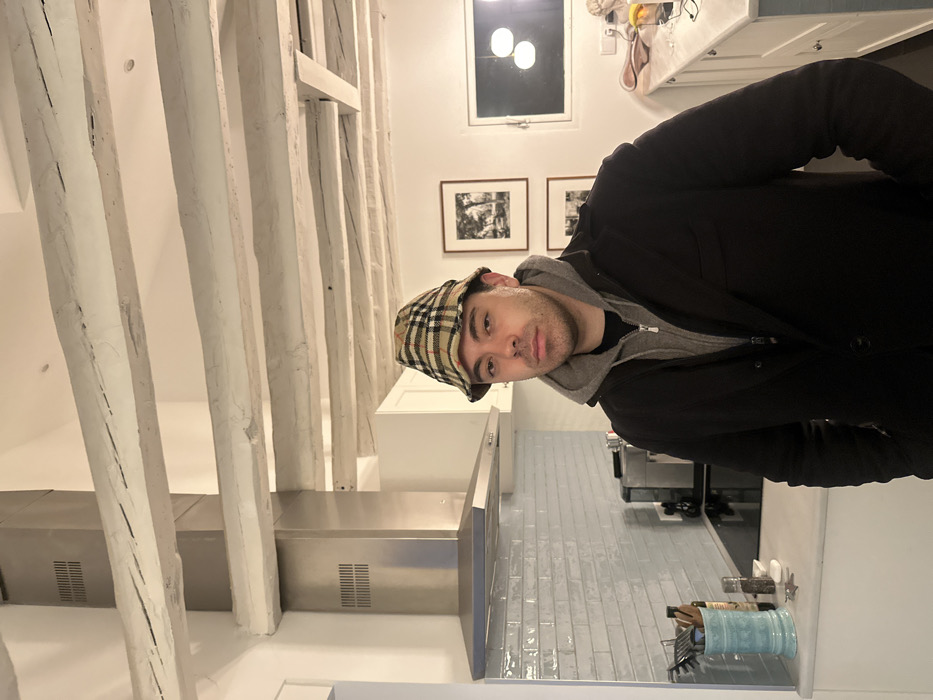 A man in a Burberry hat and dark coat stands in a kitchen. White wooden beams run along the ceiling. The kitchen has white cabinets, stainless steel appliances, and light blue backsplash tiles. Two framed black and white pictures hang on the wall. There is a window above the sink with two lights. Utensils are visible in a blue container on the counter.