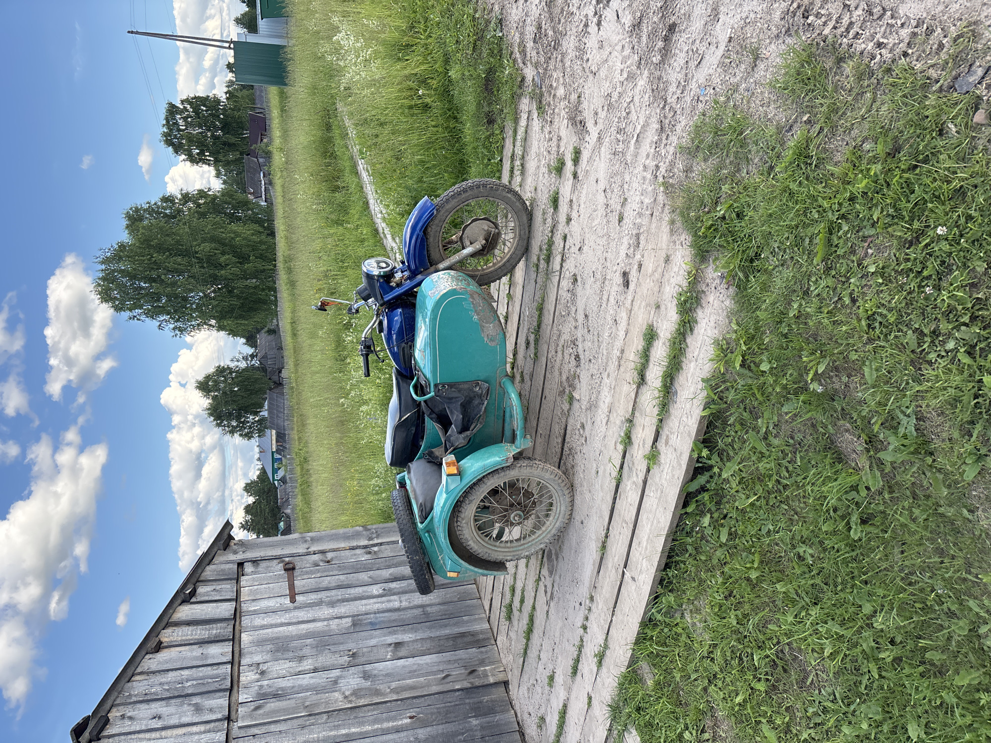 An aged, weathered wooden shed stands to the left of a dirt path, its planks vertically aligned. Next to the shed, on a raised wooden platform, is a vintage motorcycle with a sidecar. The motorcycle is primarily blue, with a turquoise sidecar. A spare tire is mounted on the rear of the sidecar. The motorcycle and sidecar appear to be in a rural setting, with a field of tall green grass stretching into the background. In the distance, several trees and houses are visible, suggesting a village or small town. The sky above is a clear blue with scattered white clouds. The scene depicts a quiet, rustic environment, possibly in or around the city of Kurgan, Russia, given the architectural style and vegetation. There are no people visible in the image, and no apparent actions are taking place.