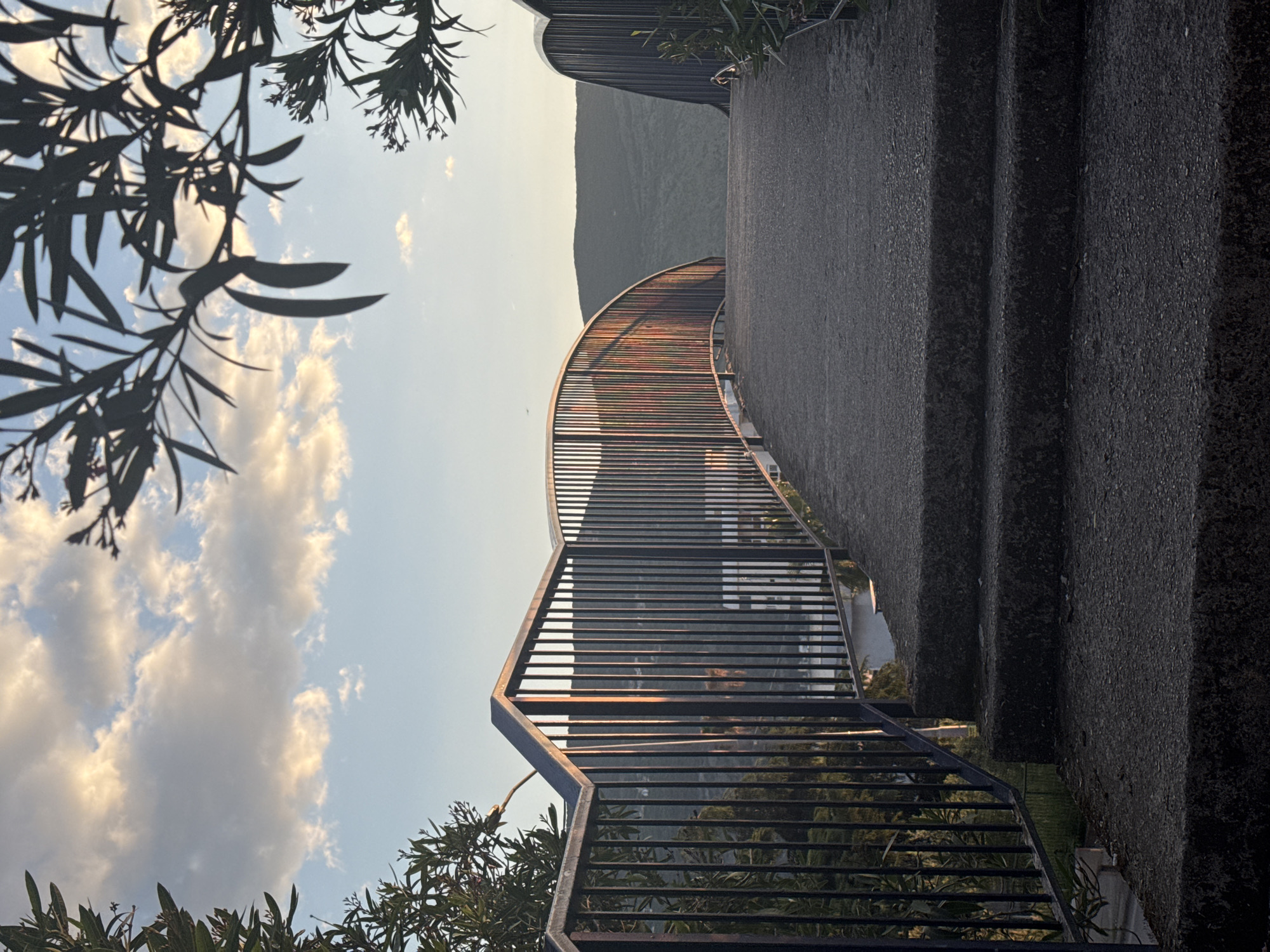 The image shows an outdoor staircase with metal railings, likely in Herceg Novi, Montenegro. The stairs are made of dark gray stone and lead upwards. The metal railings are painted black and feature horizontal bars. In the background, a hazy mountain range is visible under a light blue sky with scattered white clouds. Trees and foliage frame the image in the foreground, both at the top and bottom of the shot. The photograph is taken from a low angle, looking up the staircase.