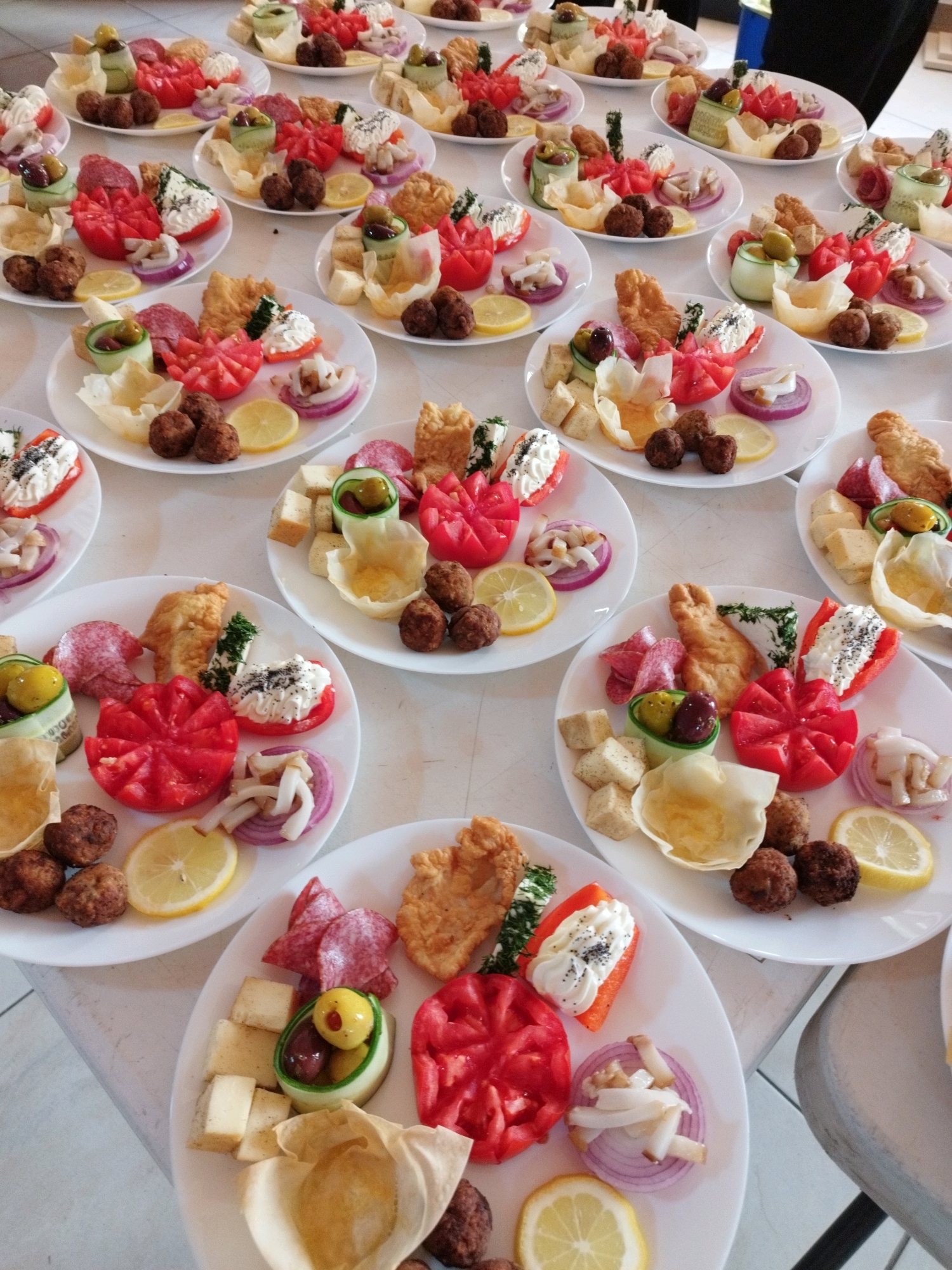 The image depicts a close-up, overhead view of numerous small plates arranged on a white surface, likely a table, for a buffet or event. Each plate contains a variety of appetizers, including sliced tomatoes, meatballs, olives, cheese cubes, salami, and some form of fried dough or pastry. There are also garnishes like lemon slices and red onion. The food is artfully arranged, suggesting it is for a catered gathering. The lighting is bright and appears to be natural or from overhead fixtures. The background is blurred, but the overall impression is of an indoor setting with a focus on the food presentation. The presence of multiple identical plates suggests a communal dining event. There are no discernible landmarks or features that would pinpoint the specific location within Bucharest, Romania. The scene is static and focuses solely on the prepared food.