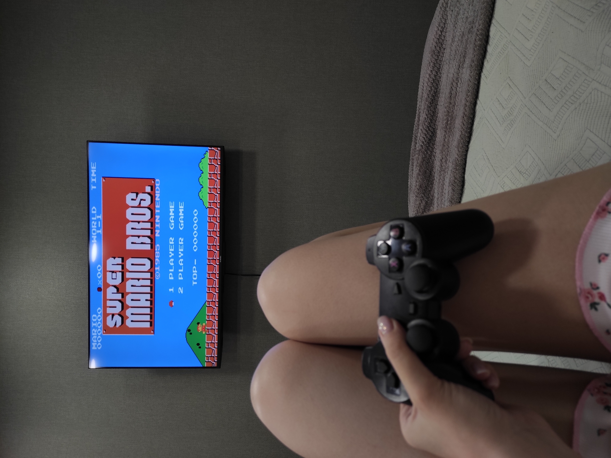 A close-up shot shows a person playing "Super Mario Bros." on a television. The person is holding a black PlayStation controller. The television is mounted on a gray wall. The person's legs are visible, positioned between the television and the edge of a patterned sofa or bed. The image does not suggest any information about location in the city of Chaikovsky, Russia.