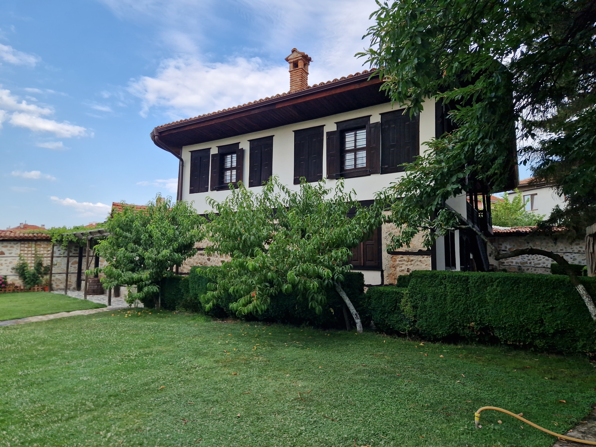 The image captures a tranquil scene of a traditional Bulgarian house in Panagyurishte, Bulgaria. The house, characterized by its white facade, dark wooden shutters, and terracotta tiled roof, stands under a bright blue sky dotted with fluffy white clouds. A red brick chimney with an ornate top stands out against the roofline. Lush green trees and neatly trimmed hedges surround the property, adding to the serene atmosphere. In the foreground, a well-maintained lawn stretches out, with a yellow garden hose visible on the right side. To the left, a cobblestone path leads to an annex building with a stone wall and a wooden pergola covered in green foliage. The overall impression is one of peaceful rural charm.