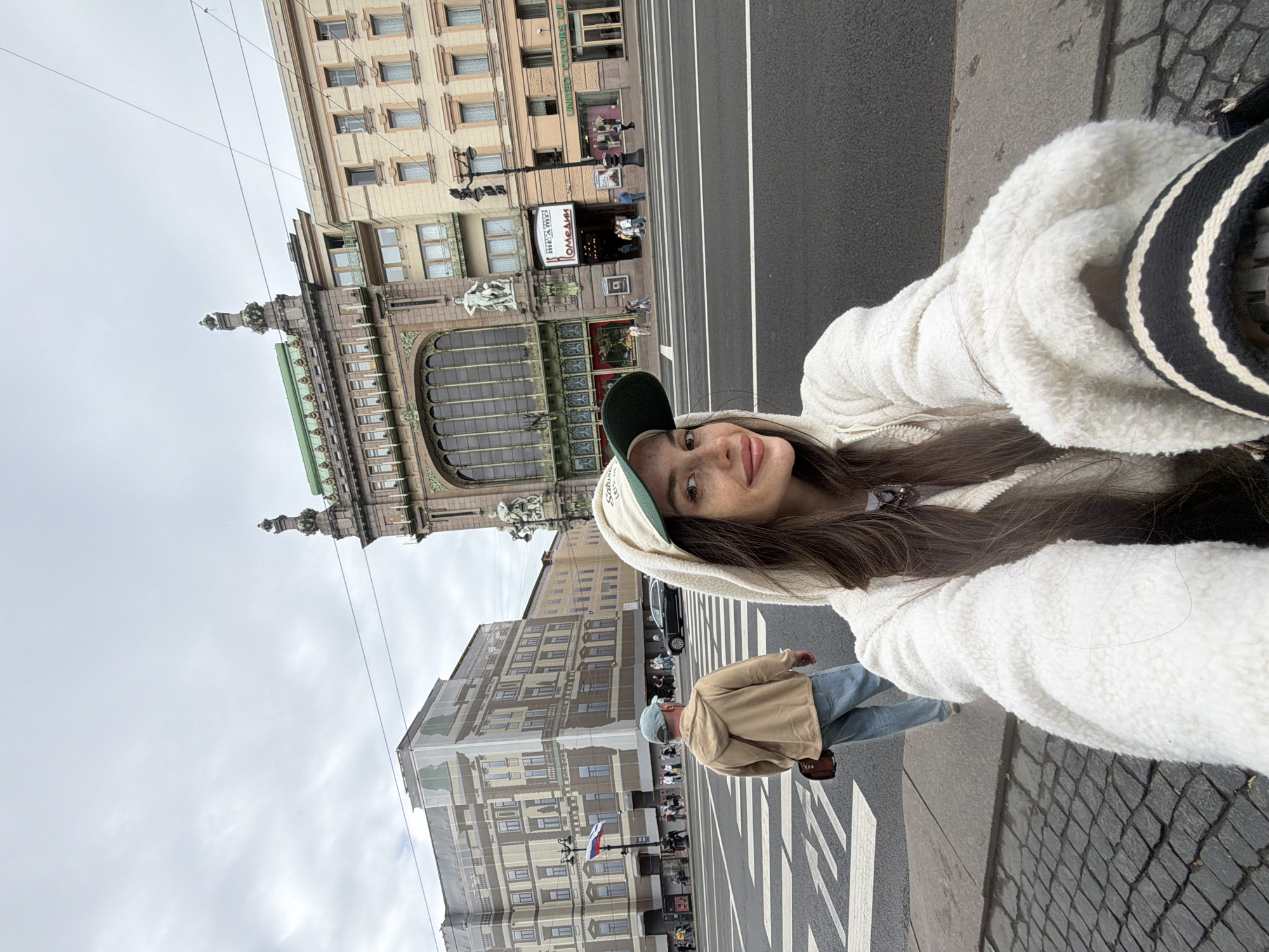 A selfie shows a young woman with long brown hair, wearing a white sherpa jacket and a green and beige baseball cap. She is standing on a sidewalk in front of a street and a large building. In the background, a man in a tan hoodie and blue jeans is walking away from the camera. The building behind them is ornate, with classical architectural elements, large arched windows, and statues. The street appears to be a wide boulevard in a city. Based on the architecture, specifically the Singer House (Dom Knigi) which is identifiable by its distinctive dome and facade, this scene is located on Nevsky Prospekt in Saint Petersburg, Russia. The sky is overcast.