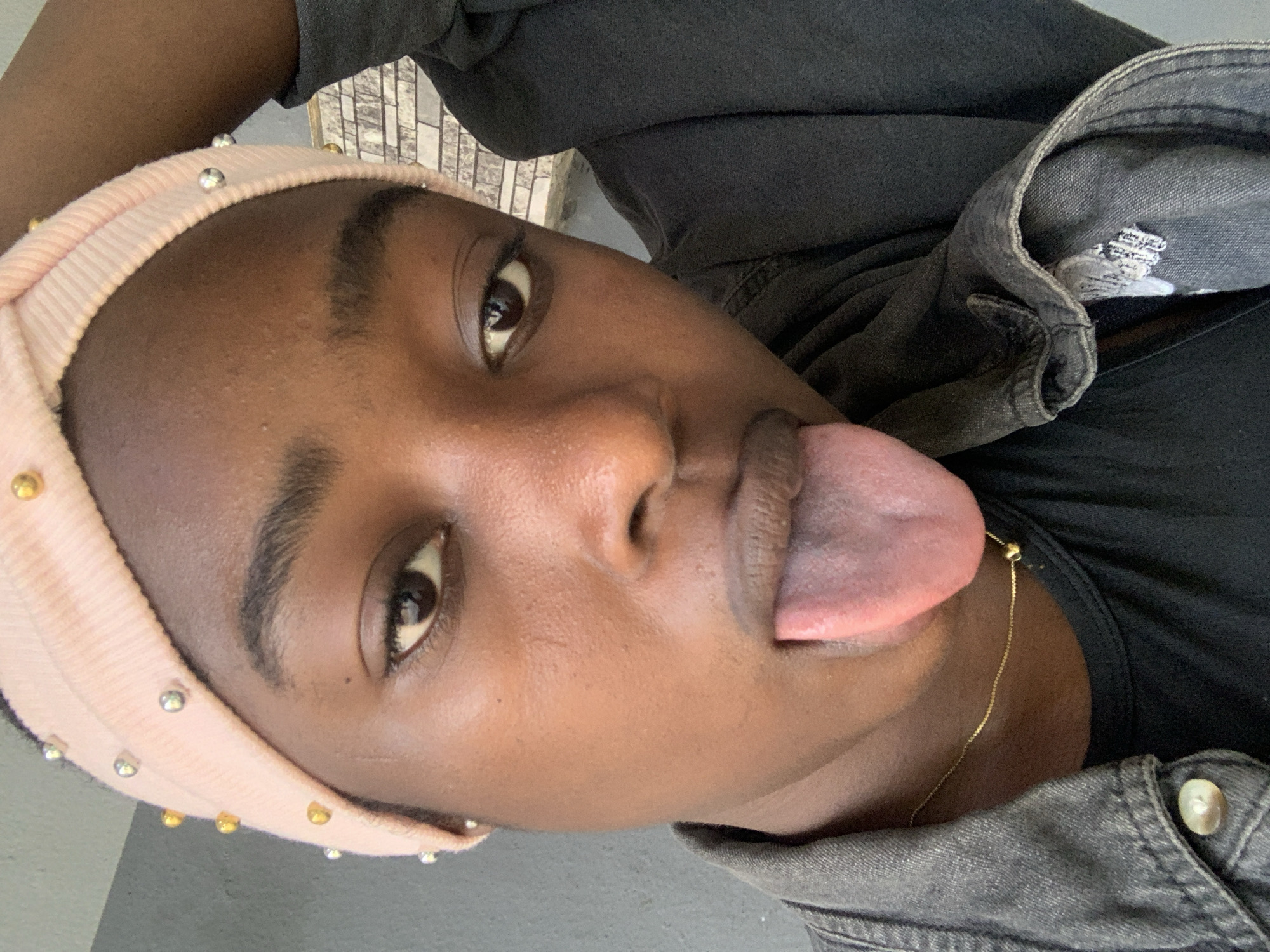 A close-up, rotated shot of a dark-skinned person with a pink beaded headband sticking their tongue out. They are wearing a black shirt and a denim jacket. A gold chain necklace is visible. The background appears to be a light grey, possibly a floor. There is no indication of the location being New Stadium, Nigeria in the image.
