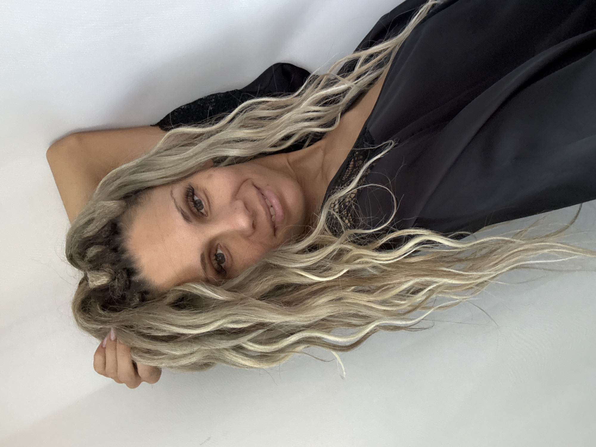 A woman is captured in a portrait-style image, leaning against a light-colored wall. Her head is tilted back, with her right arm bent and resting behind her head, her hand touching her hair. Her long, blonde, wavy hair cascades down. She is wearing a black satin robe with lace trim on the neckline. Her expression is a gentle smile, with her eyes looking towards the camera. The lighting is soft, and the background is a plain white wall, suggesting an indoor setting. There is no information in the image to determine the location within the city of Rusanovo, Russia.