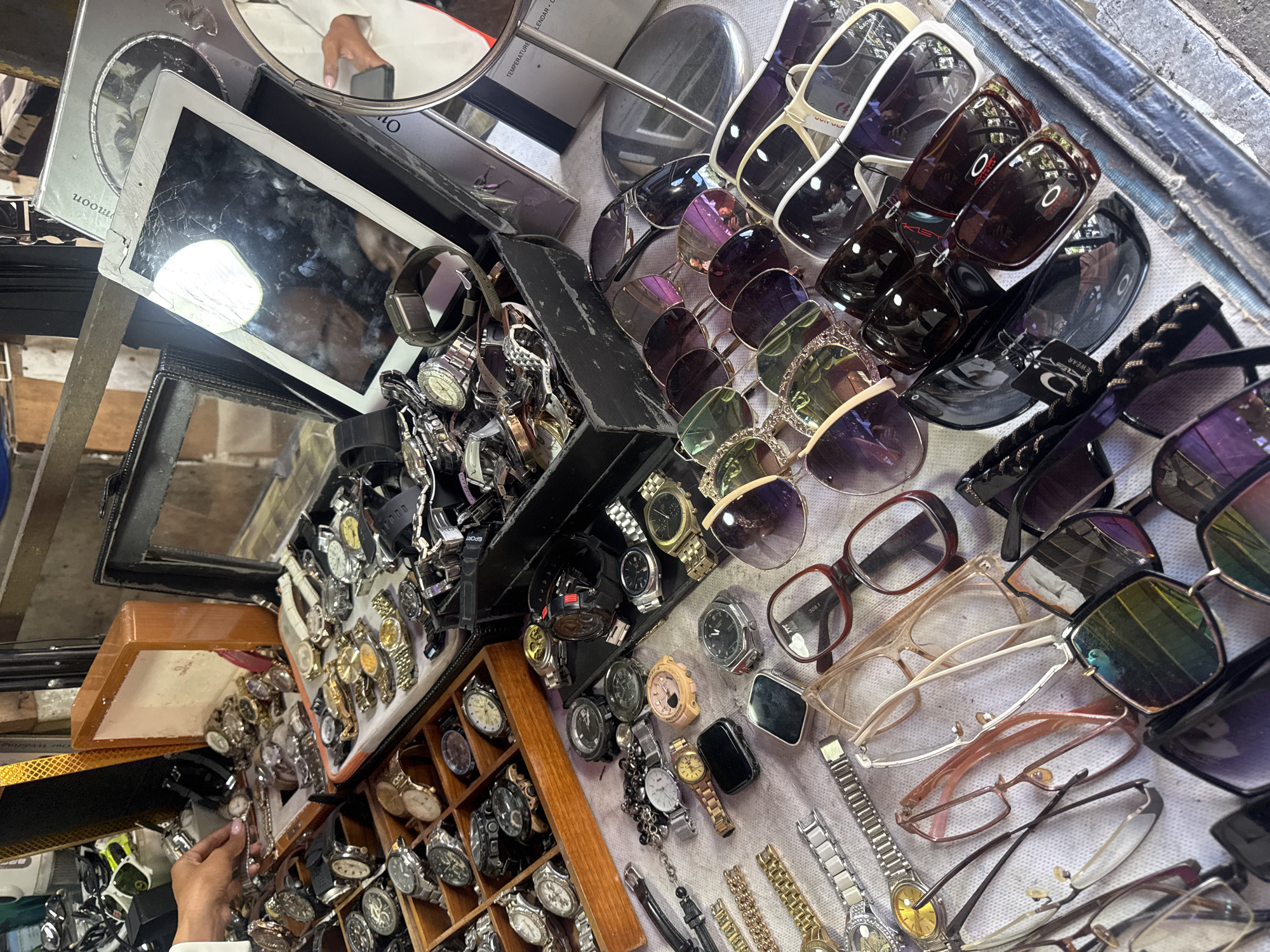 An overhead, medium shot captures a street market stall in Denpasar, Indonesia, displaying a variety of wristwatches and sunglasses. The stall is cluttered, with numerous watches arranged in wooden boxes, on fabric surfaces, and in black plastic containers. Some watches have metal bands, while others have leather or rubber straps. Several pairs of sunglasses are also visible, with different frame styles and colored lenses. In the background, a cracked tablet screen is propped up, and other reflective surfaces and metallic objects are present. A hand, presumably belonging to a vendor or customer, is reaching into one of the watch boxes. The overall impression is of a bustling, open-air market with a wide assortment of accessories.
