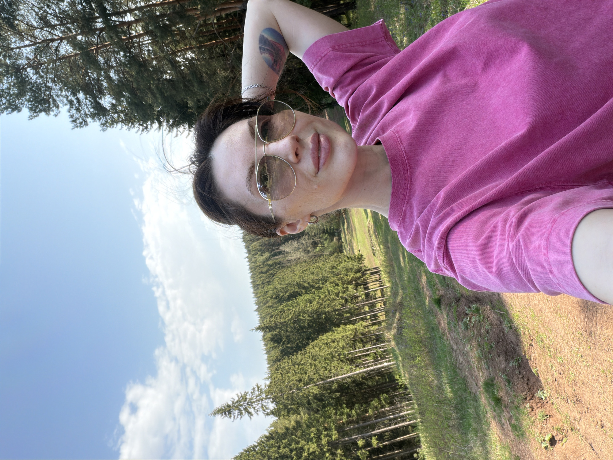 A woman is taking a selfie outdoors. She is wearing a pink t-shirt and round sunglasses. Her left arm is raised, displaying a tattoo. A forest is in the background, with a blue sky and white clouds visible above the trees. The ground is covered in grass and dirt. The location is likely a forested area outside the city center.