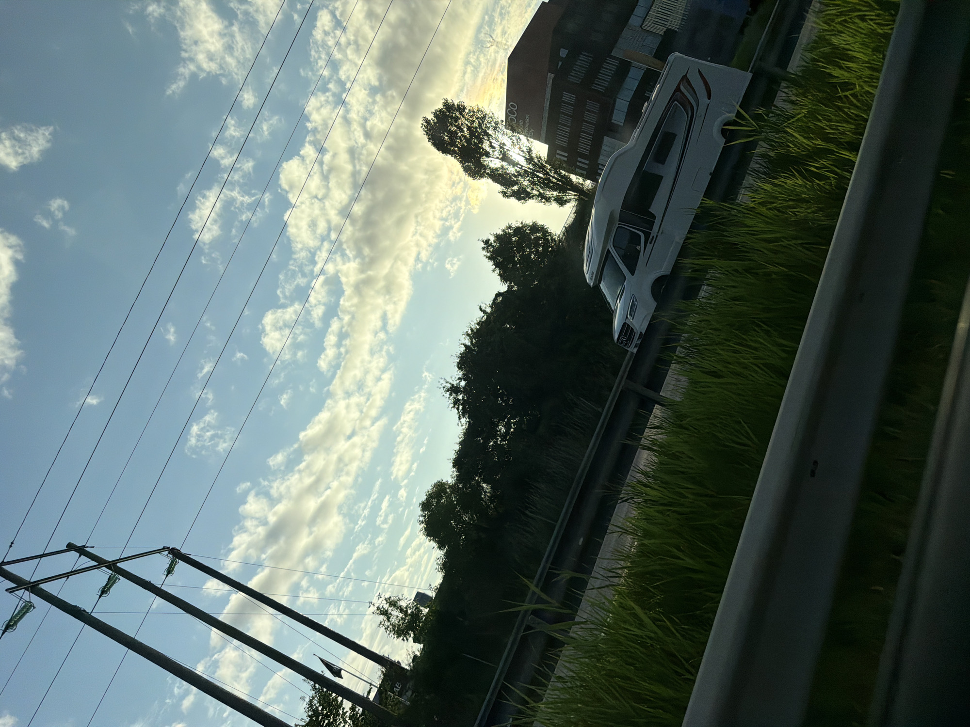 The image depicts a white camper van driving on a highway in Mölndal, Sweden, likely during late afternoon or early evening. Tall power lines are visible in the sky, with multiple wires suspended between two large wooden poles. The sky is a light blue, with scattered white and yellowish clouds, suggesting the sun is low on the horizon.
In the foreground, lush green grass grows alongside the highway, separated by a metal guardrail. Beyond the highway, a dense line of trees forms a dark green barrier. To the right of the frame, a modern brick building with large windows is visible, bearing the text "COCO" on its facade, indicating it's part of a business or innovation park. A tall, slender tree stands in front of the building, silhouetted against the brighter sky. The camper van is the primary subject in motion, traveling on the highway. No people are clearly visible within or around the van. The overall scene suggests a journey through an urban or suburban area with interspersed natural elements.