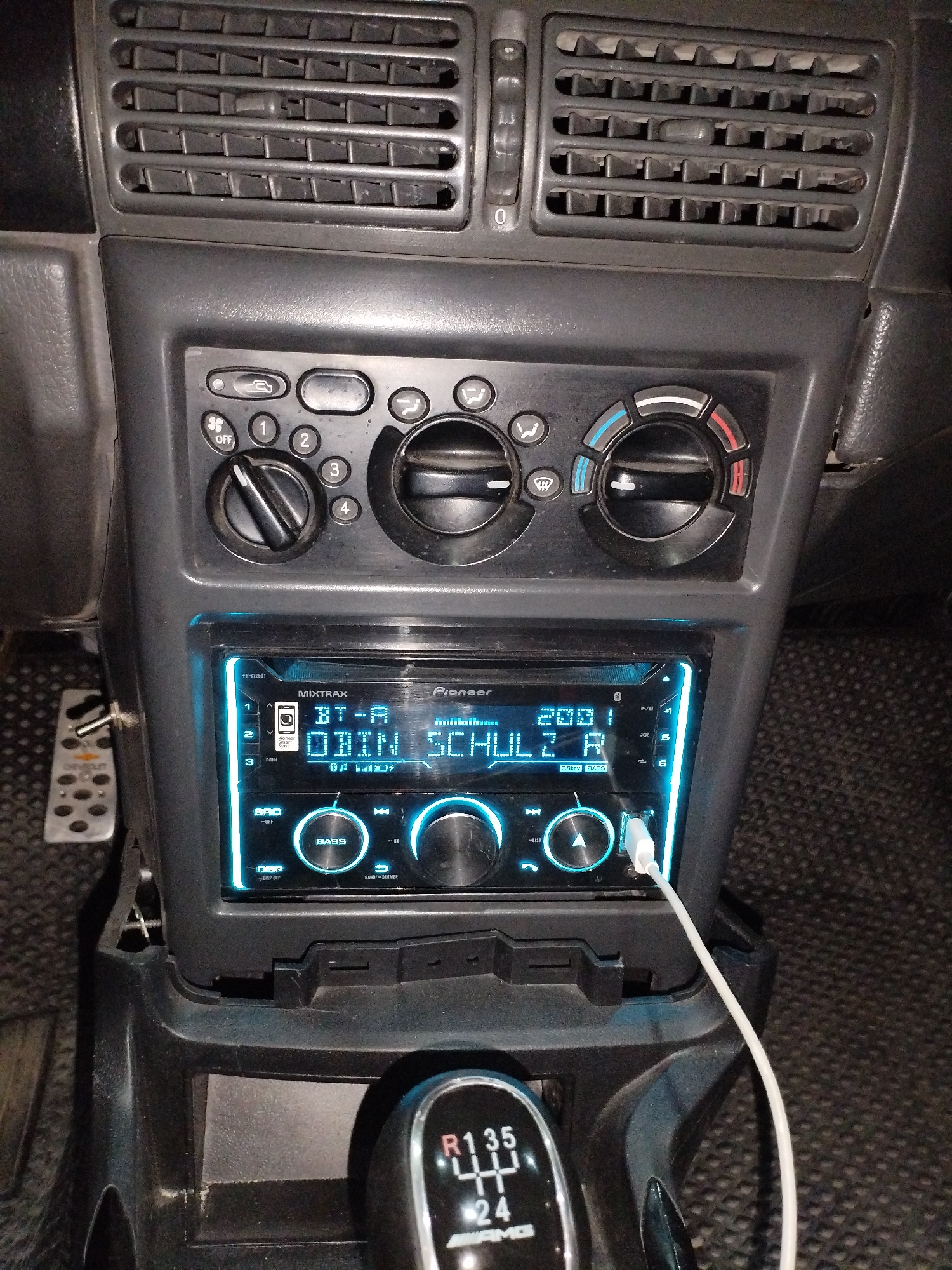 A close-up shot captures the interior of a car, focusing on the dashboard and center console. The dashboard features air vents at the top, followed by climate control knobs and buttons. Below that is a Pioneer car stereo with a bright blue illuminated display. The screen shows "MIXTRAX" and "Pioneer" branding, along with text that reads "BT-A OBIN SCHULE R" and what appears to be a song title or artist. A white USB cable is plugged into the front of the stereo. To the left of the stereo, a foot pedal is visible. The center console has a gear shift knob with visible gear markings (R, 1, 3, 5, 2, 4) and an "AMG" logo. The overall environment appears to be dark, with a textured floor mat visible at the bottom. The scene is set within a car, likely in a parked location. There are no people visible in the image. The location of Termez, Uzbekistan is not discernible from the visual information provided in the image.