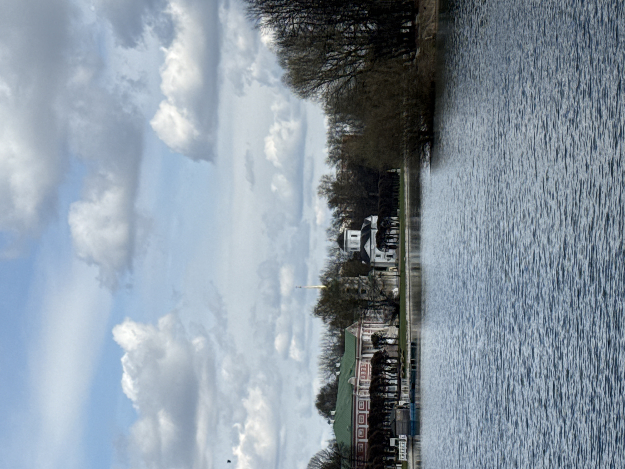 This image shows the cityscape of Moscow, Russia, situated near a body of water. A red and white building is present on the shoreline and behind it is a white church. In the sky, white clouds are visible. The image has been rotated 90 degrees.